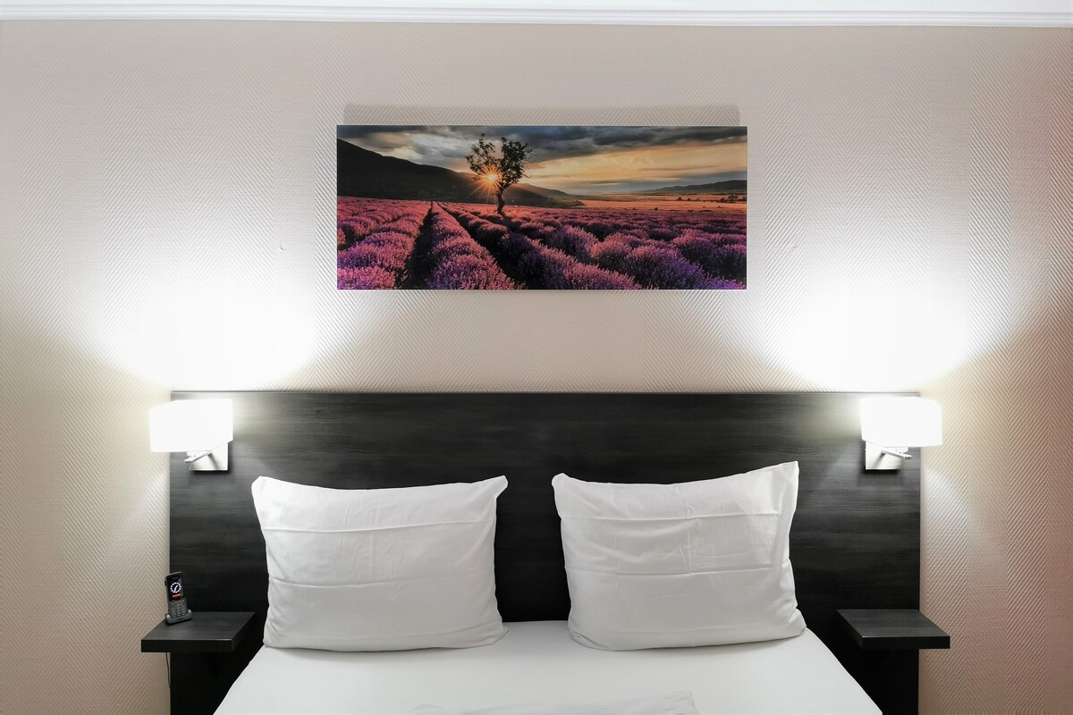 Habitación de hotel con cama, dos almohadas blancas, mesita de noche con teléfono y un cuadro en la pared de campos de lavanda al atardecer.