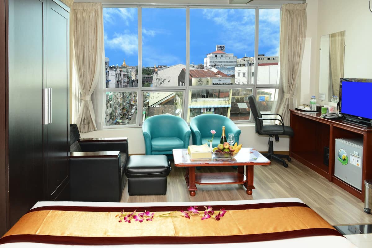 Een hotelkamer met een groot raam, een bed met een bloemenprint, twee teal fauteuils, een houten bureau met een computer, en een minibar.