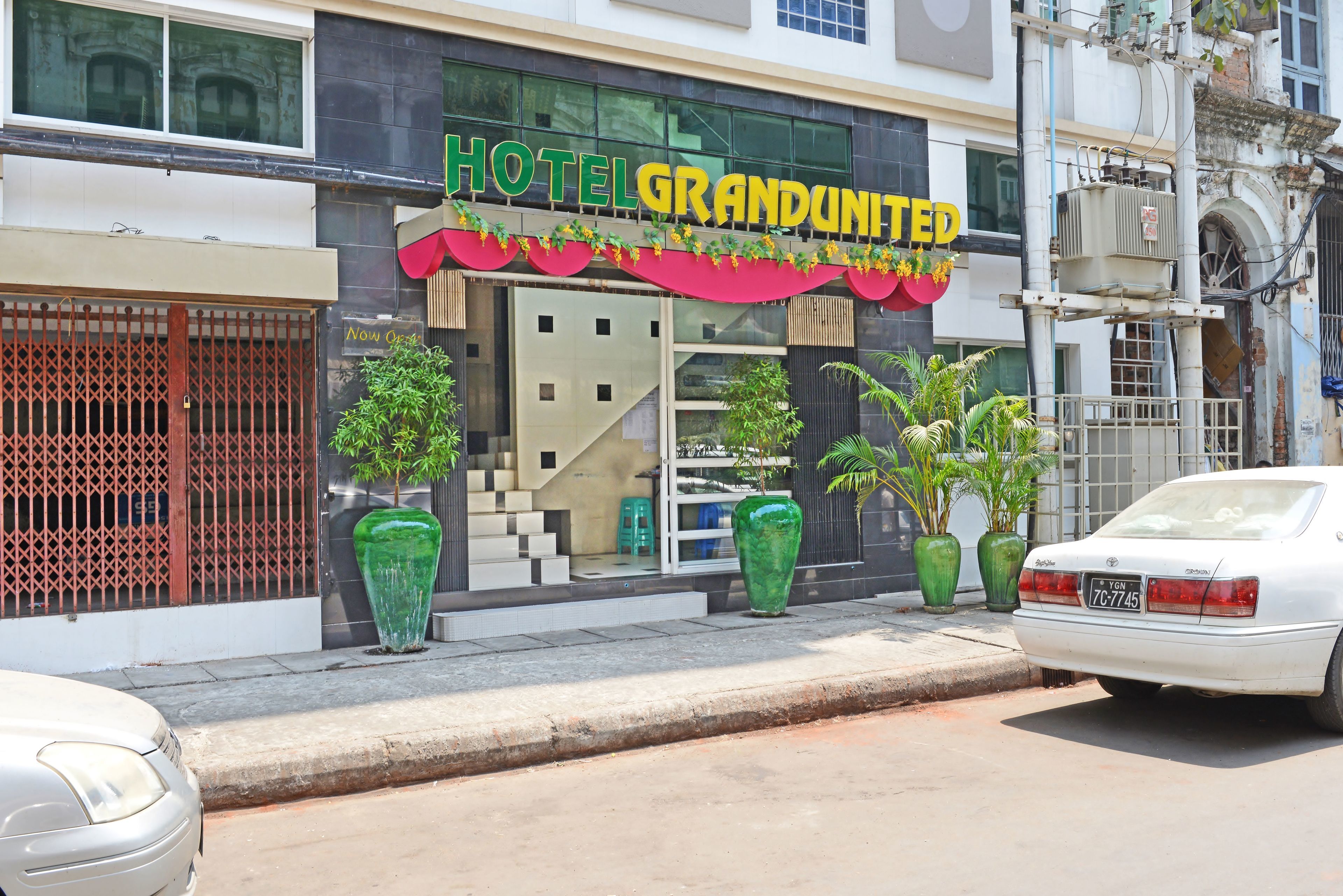 A hotel named "HOTEL GRANDUNITED" with a modern facade, large windows, and potted plants in front.
