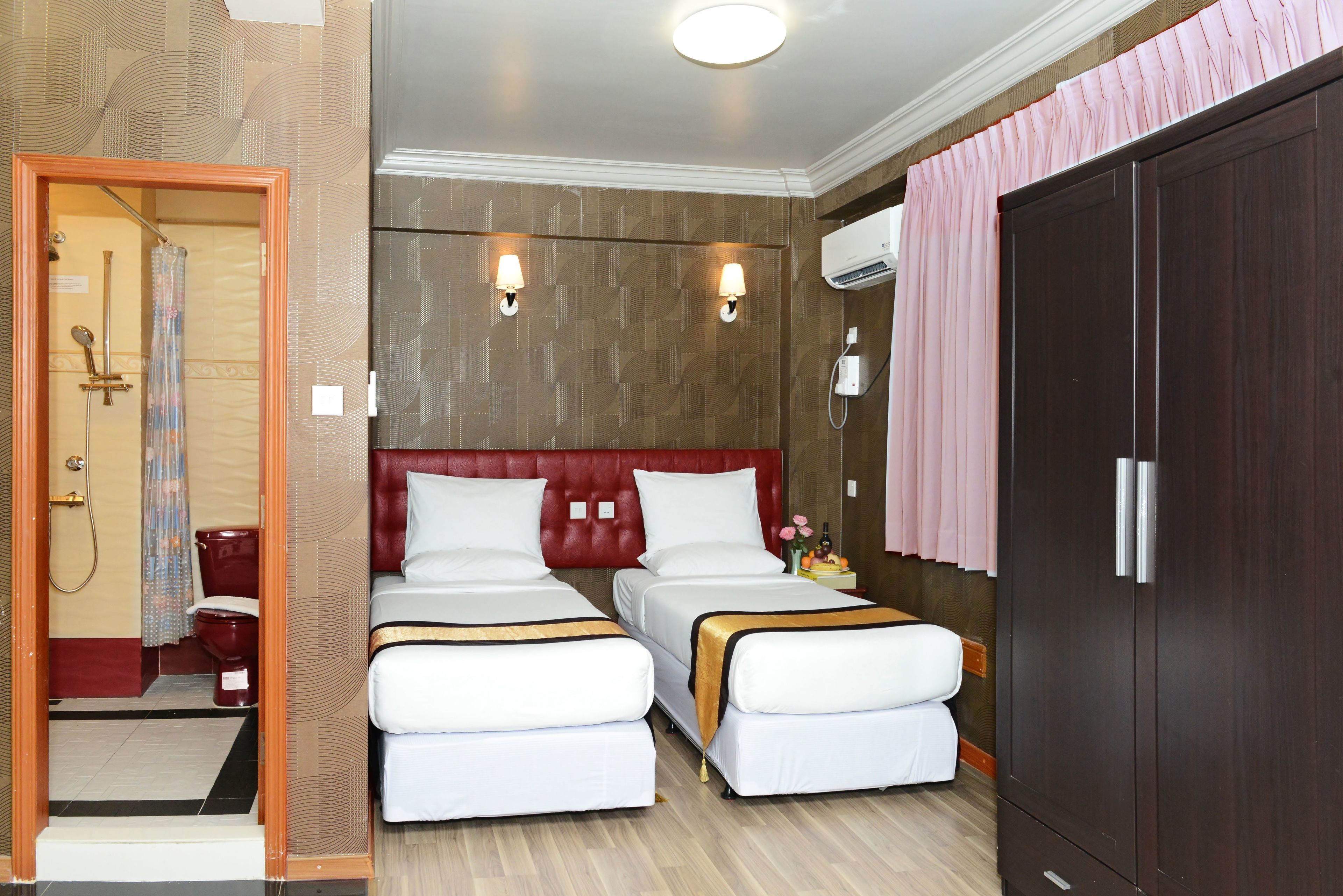 Una habitación de hotel con dos camas, cabecero de madera, armario, unidad de aire acondicionado y baño con ducha e inodoro.