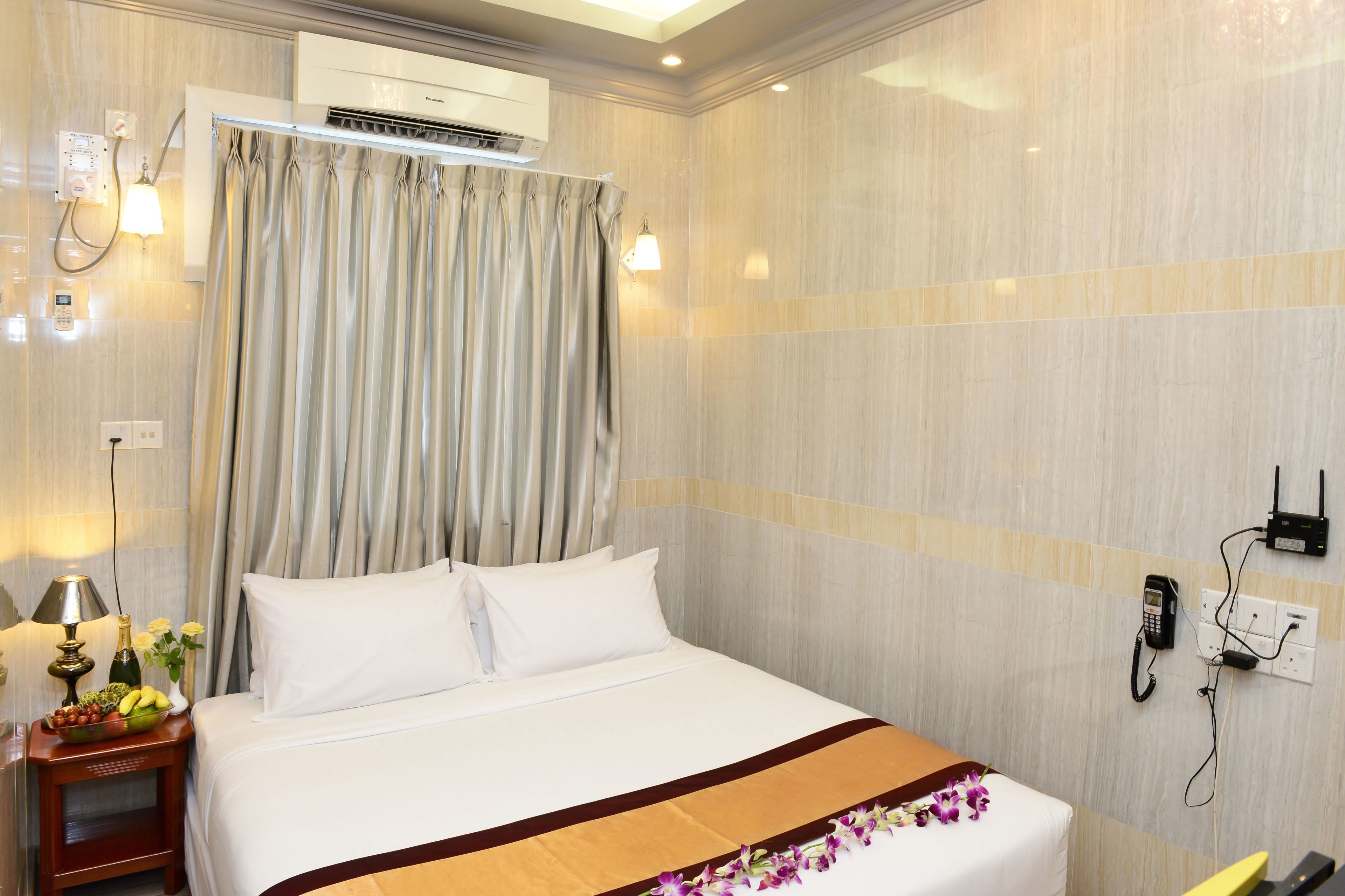 A hotel room with a bed, a nightstand with a lamp and a fruit bowl, an air conditioning unit, a telephone, and a curtain.