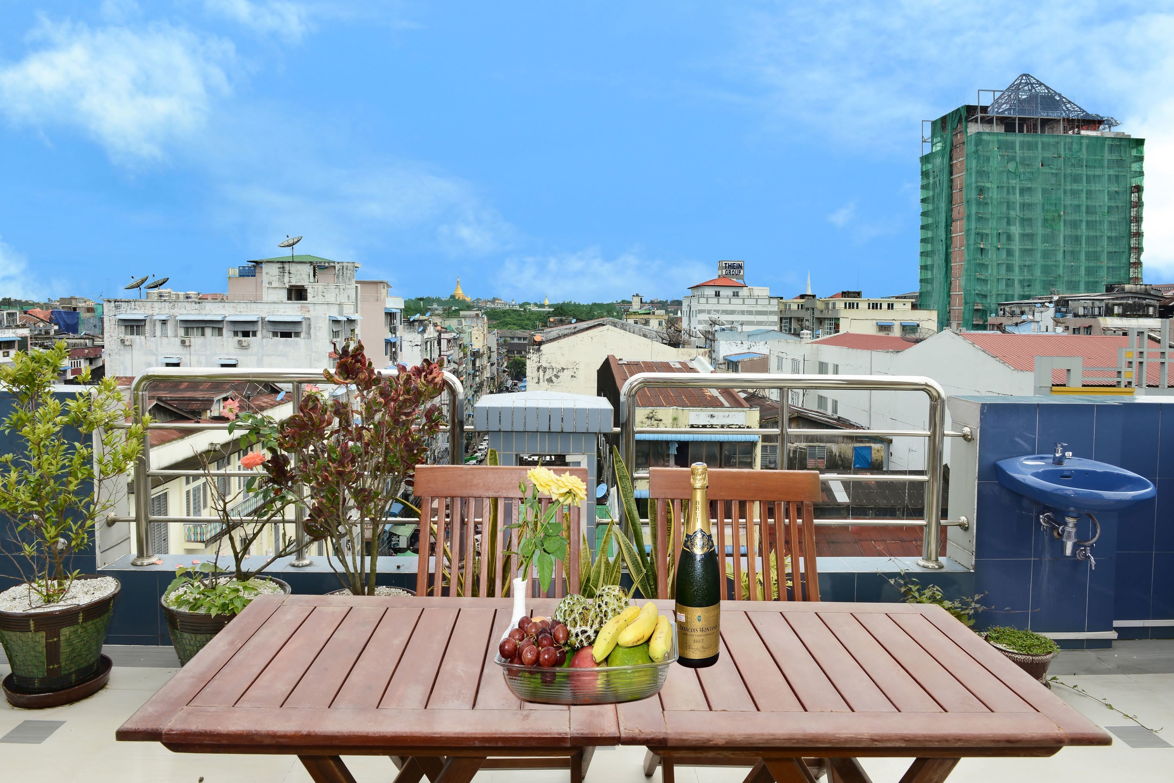 A wooden table with a fruit bowl, a bottle, and potted plants on a balcony with a city view.