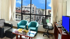 Ein Zimmer mit einem großen Fenster, zwei sesseln in Türkis, einem Holztisch mit Früchten und einer Flasche, einem schwarzen Bürostuhl und einem Schreibtisch mit einem Computermonitor.
