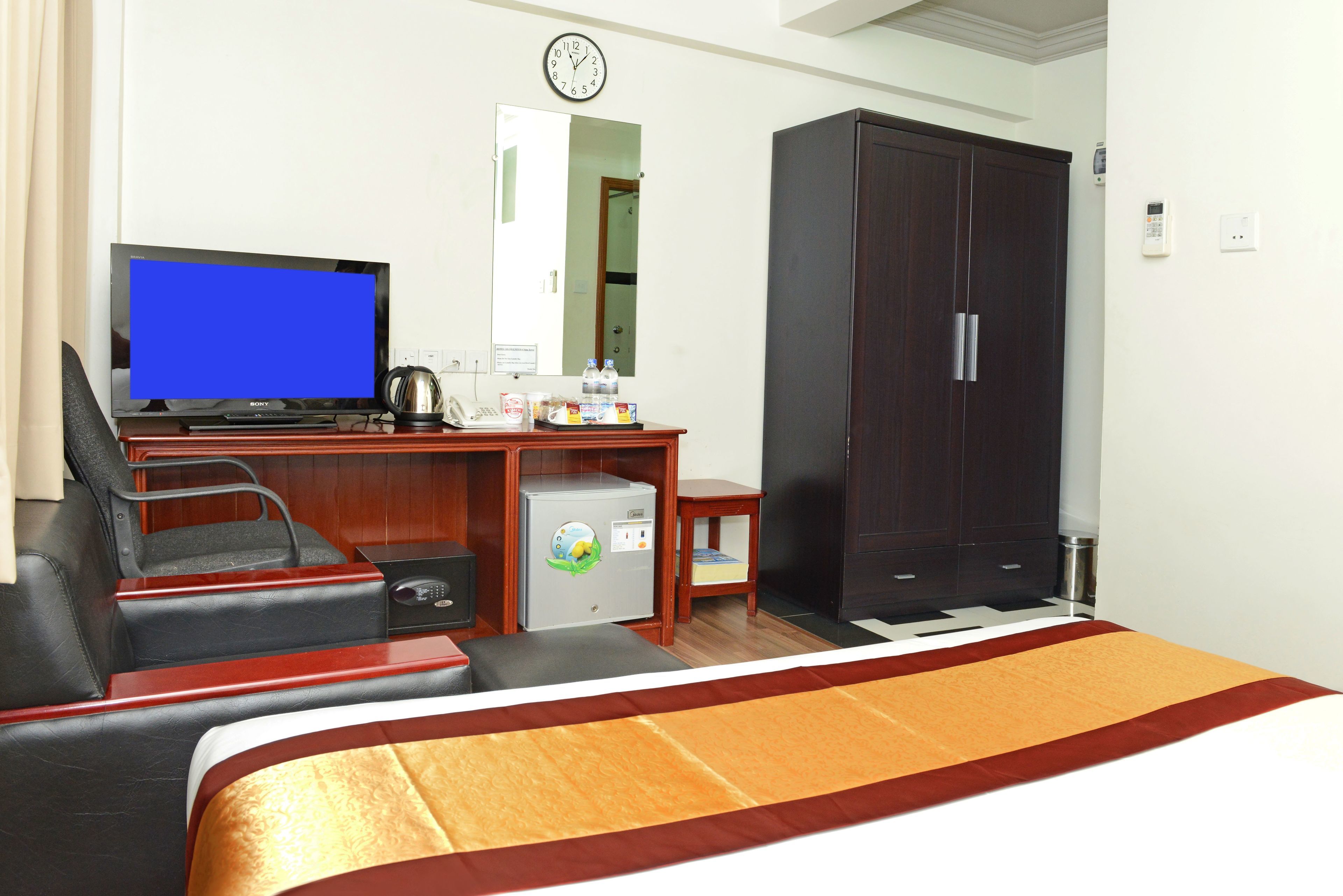 A hotel room with a bed, a desk with a computer and a chair, a wardrobe, a clock, and a small refrigerator.