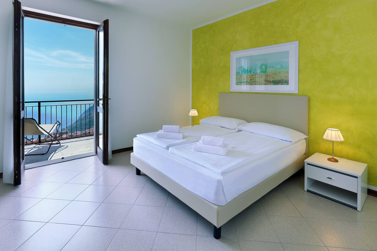 A hotel room with a bed, a nightstand, a lamp, a painting, and a balcony with a chair.
