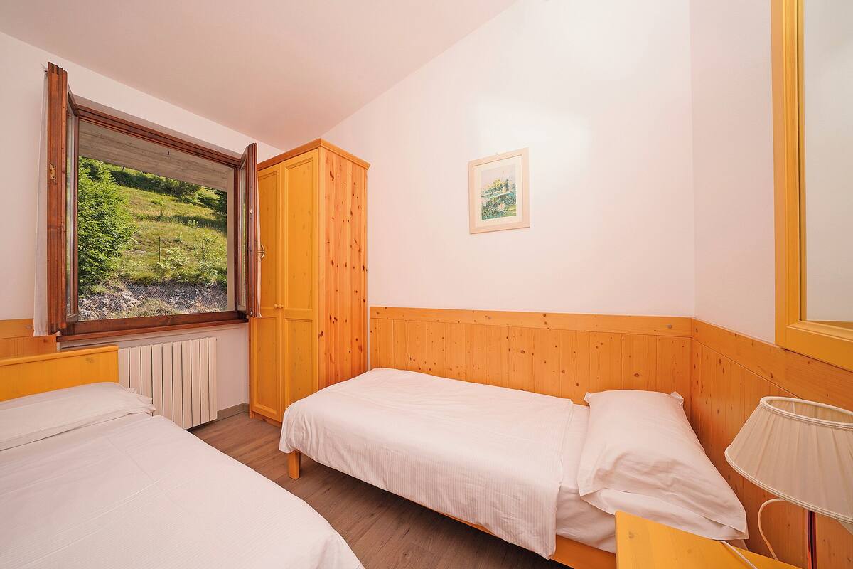 A bedroom with two beds, a wooden wardrobe, a window with a view of greenery, and a framed picture on the wall.