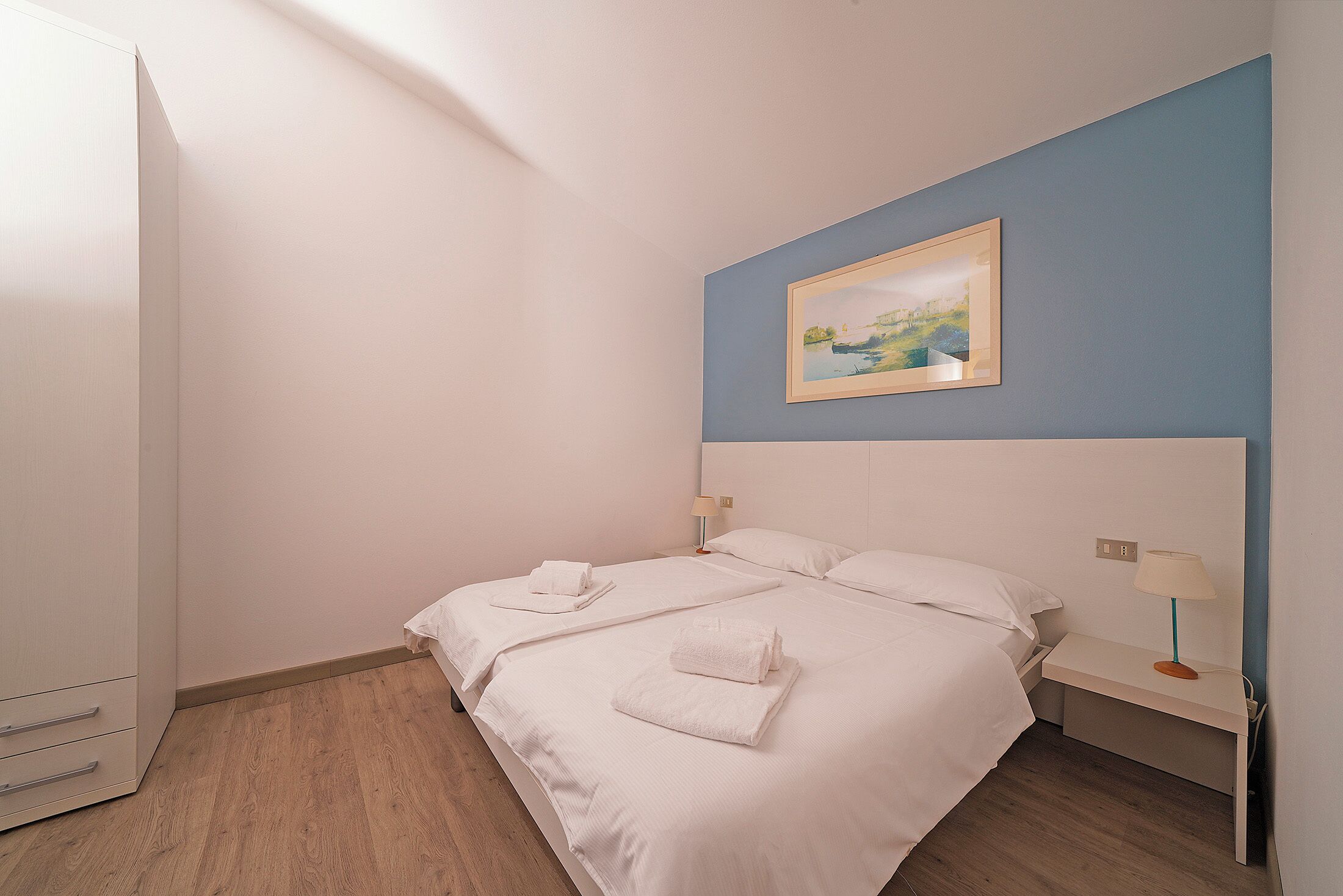 A hotel room with two beds, a painting on the wall, and a desk with a lamp.