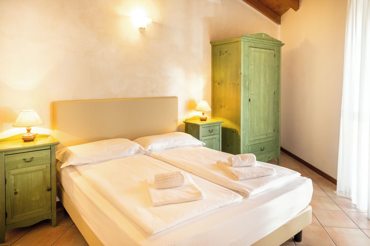 A neatly made bed with white linens and towels, a green nightstand with a lamp, and a green wardrobe.