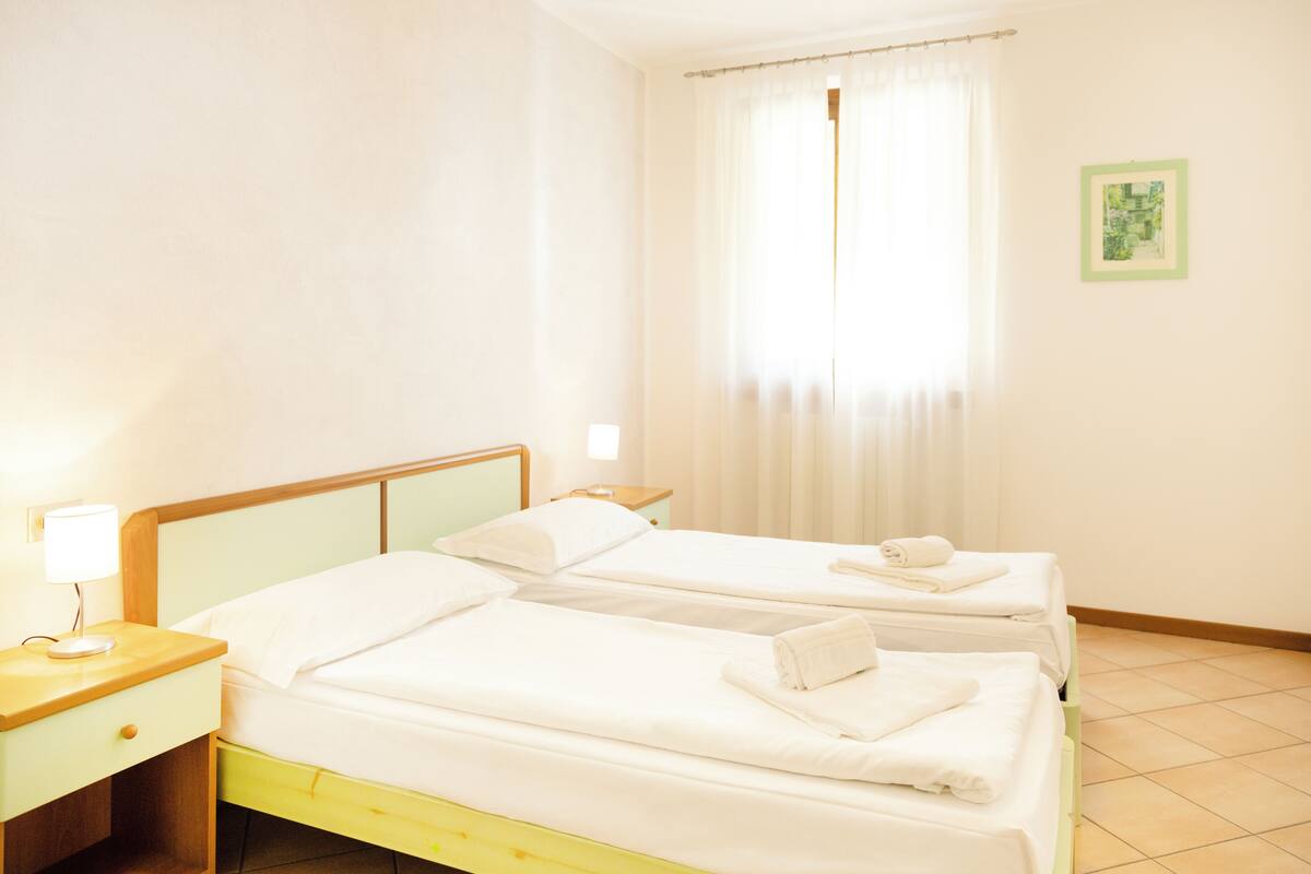 A hotel room with two beds, a nightstand with a lamp, a window with curtains, and a framed picture on the wall.