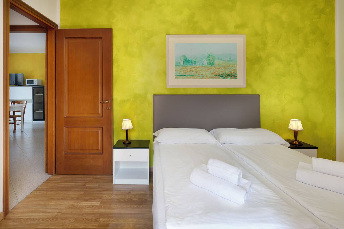 A hotel room with a bed, two bedside tables with lamps, a painting on the wall, and a door leading to a dining area.