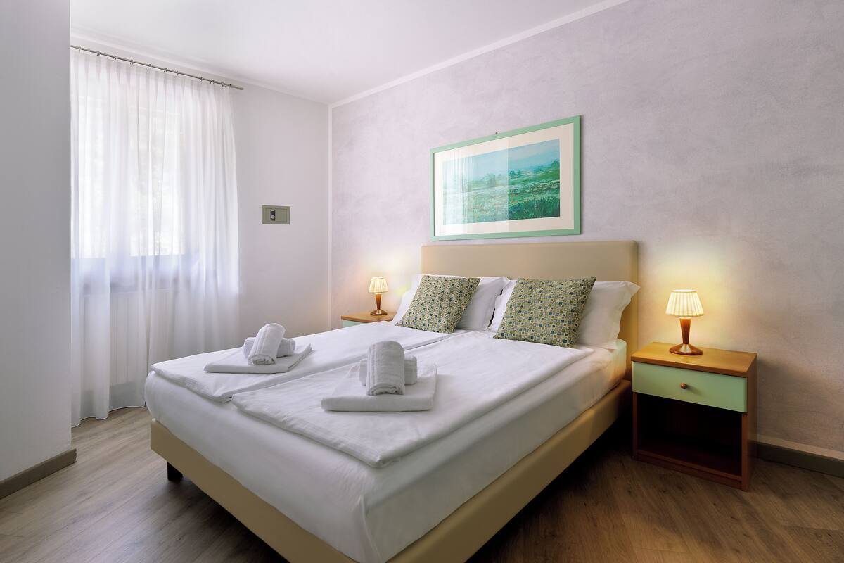 A double bed with white linens, two bedside tables with lamps, and a painting on the wall.