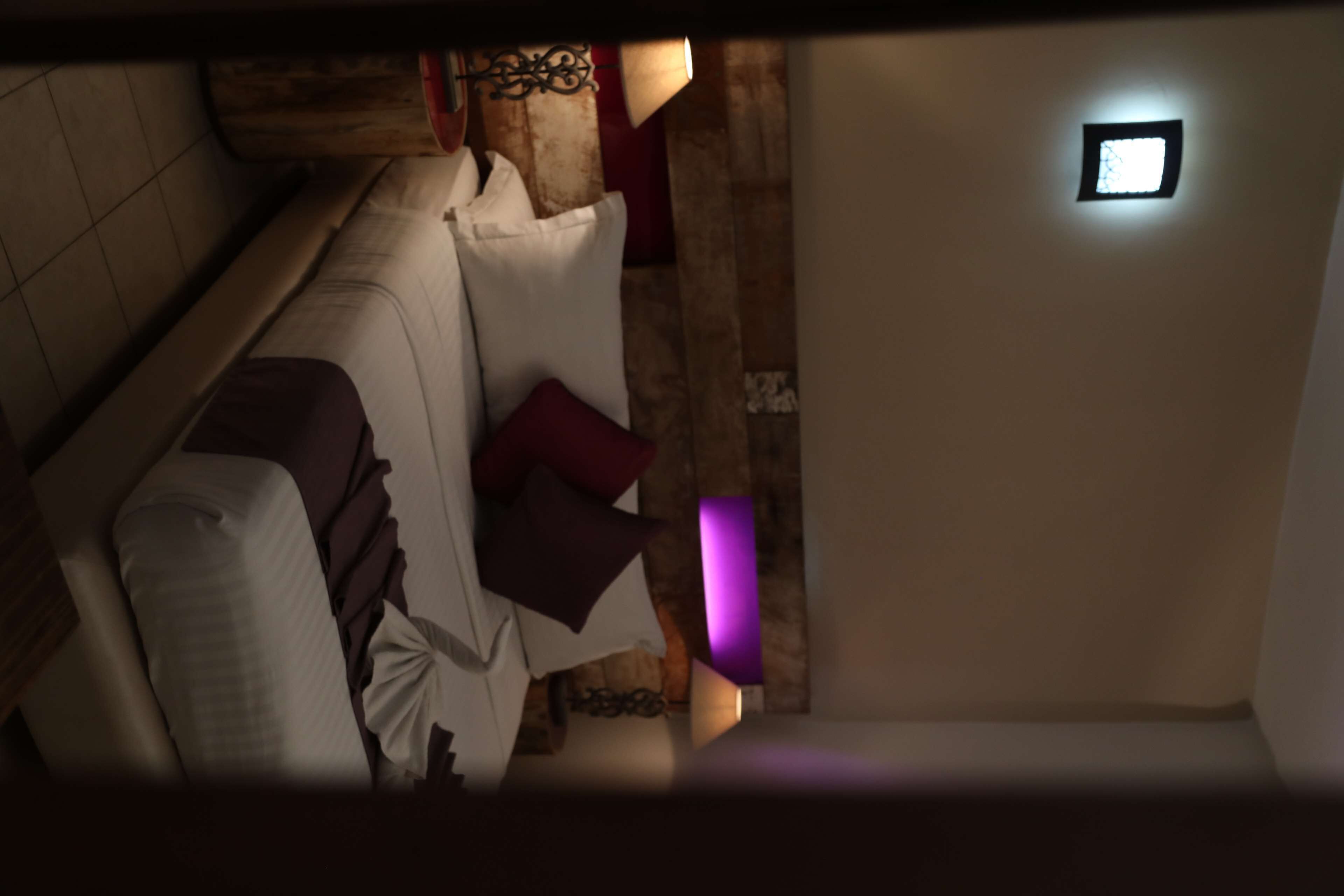 A dimly lit room with a wooden floor, a white sofa with a patterned cushion, and a small purple light fixture on the wall.