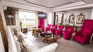 A modern living room with a white sofa, multiple pink armchairs, and round coffee tables. A flat-screen TV is mounted on the wall, and there are decorative mirrors and a fan. - The Swiss Hotel (Freetown)