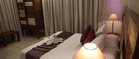 A hotel room with a bed, a bedside lamp, a wooden wardrobe, a bench, and curtains.