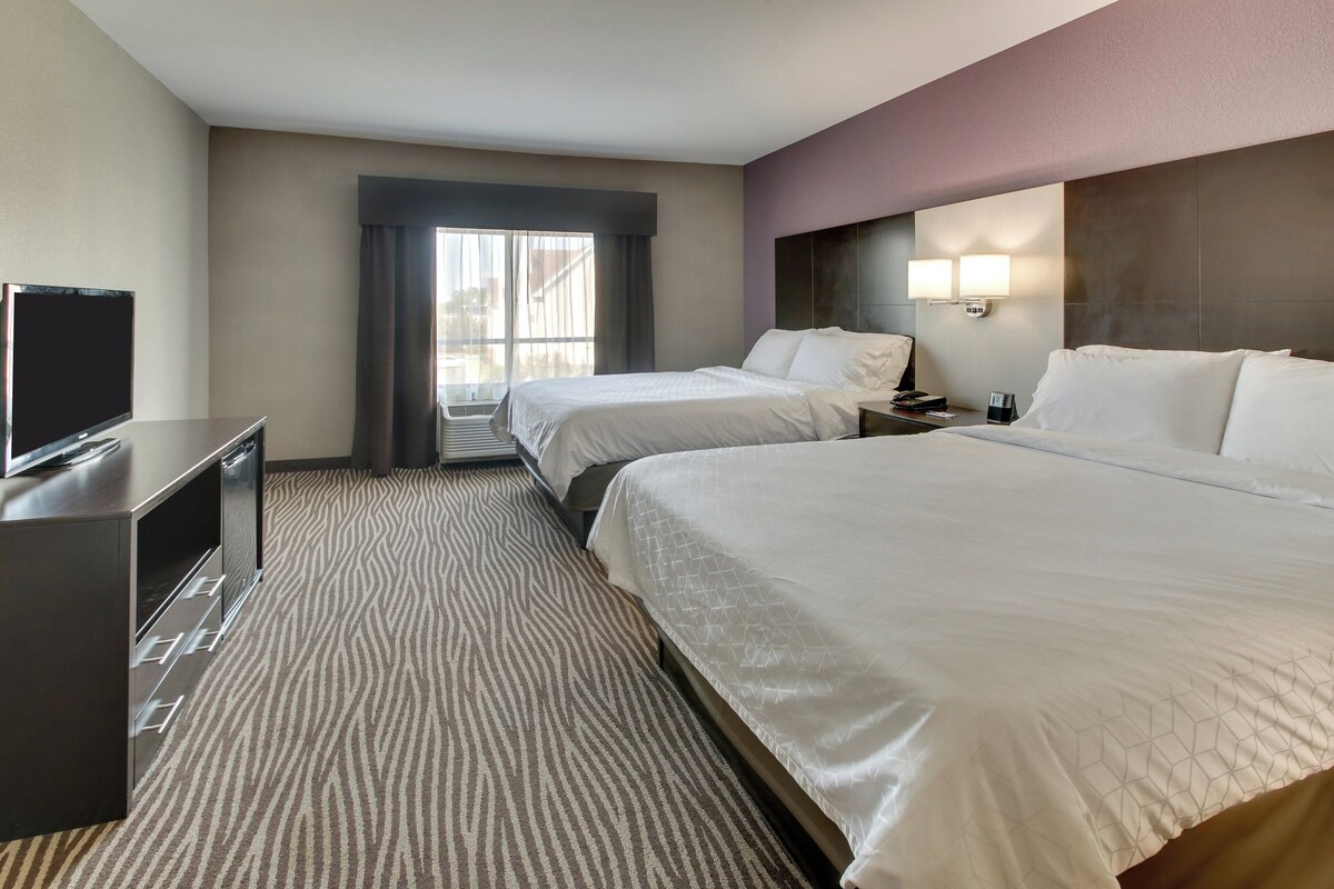 A hotel room with two beds, a TV, a desk, and a window with curtains.