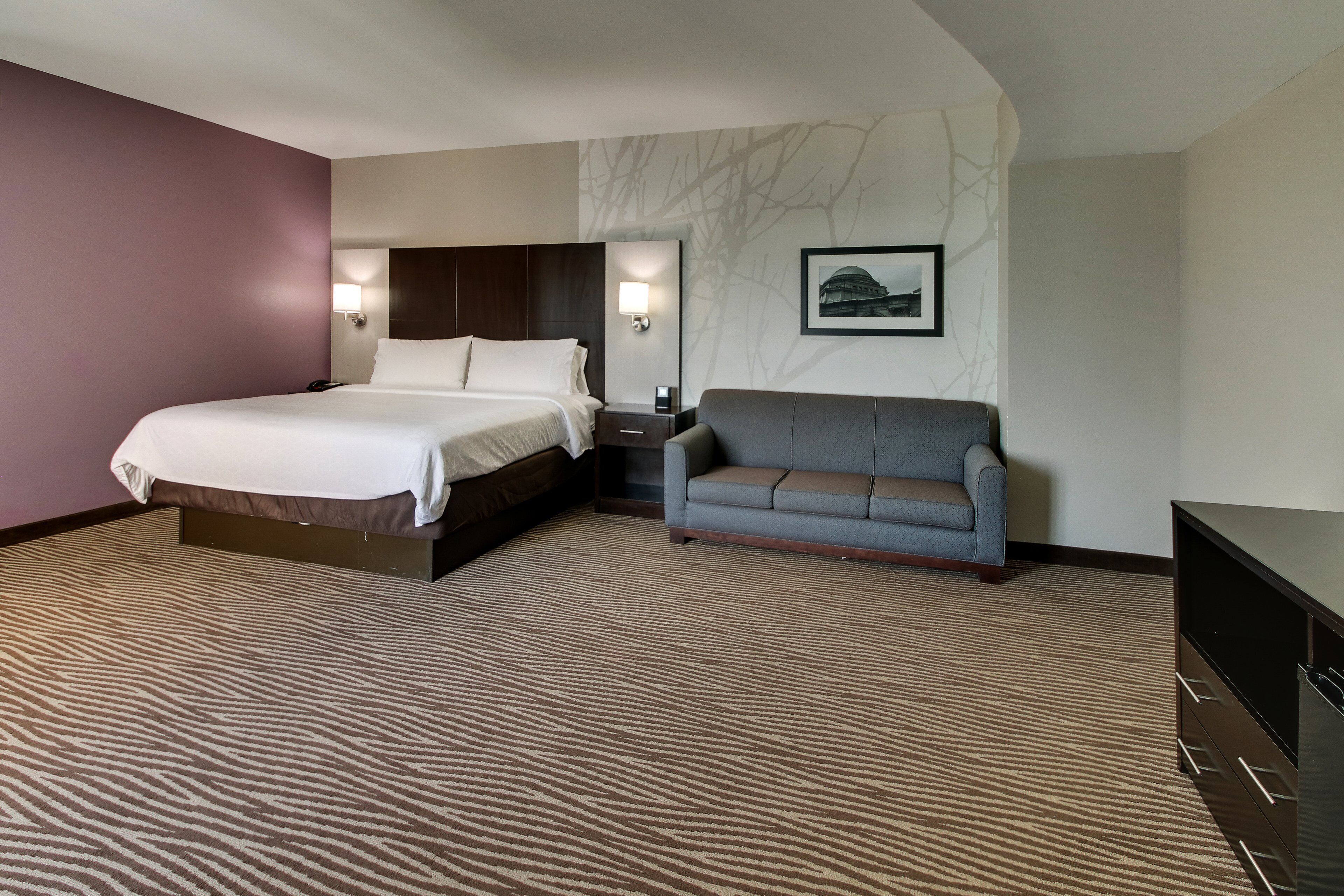 A hotel room with a bed, a sofa, a nightstand, a lamp, and a picture on the wall.