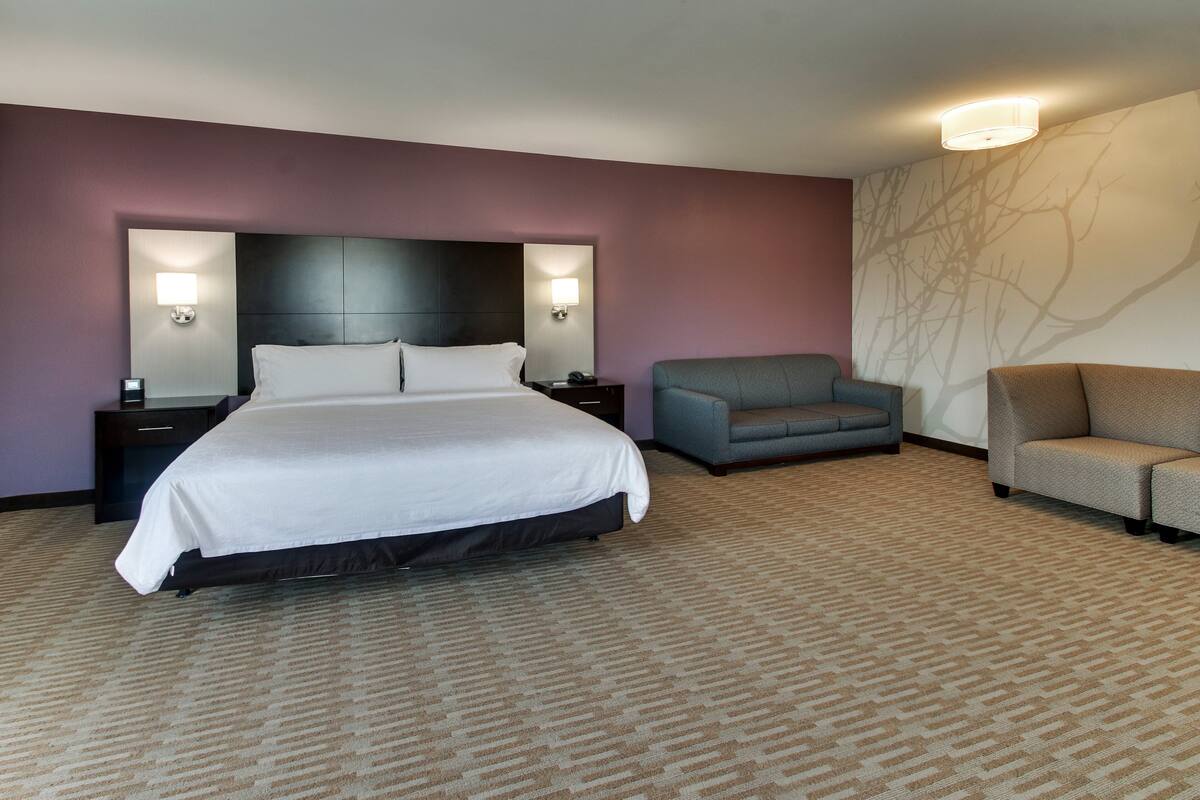 A hotel room with a large bed, two nightstands, a gray armchair, and a sofa.