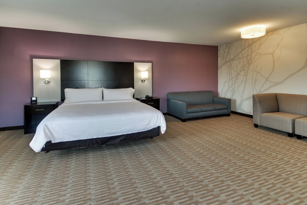 A hotel room with a large bed, two nightstands, a gray armchair, and a sofa.