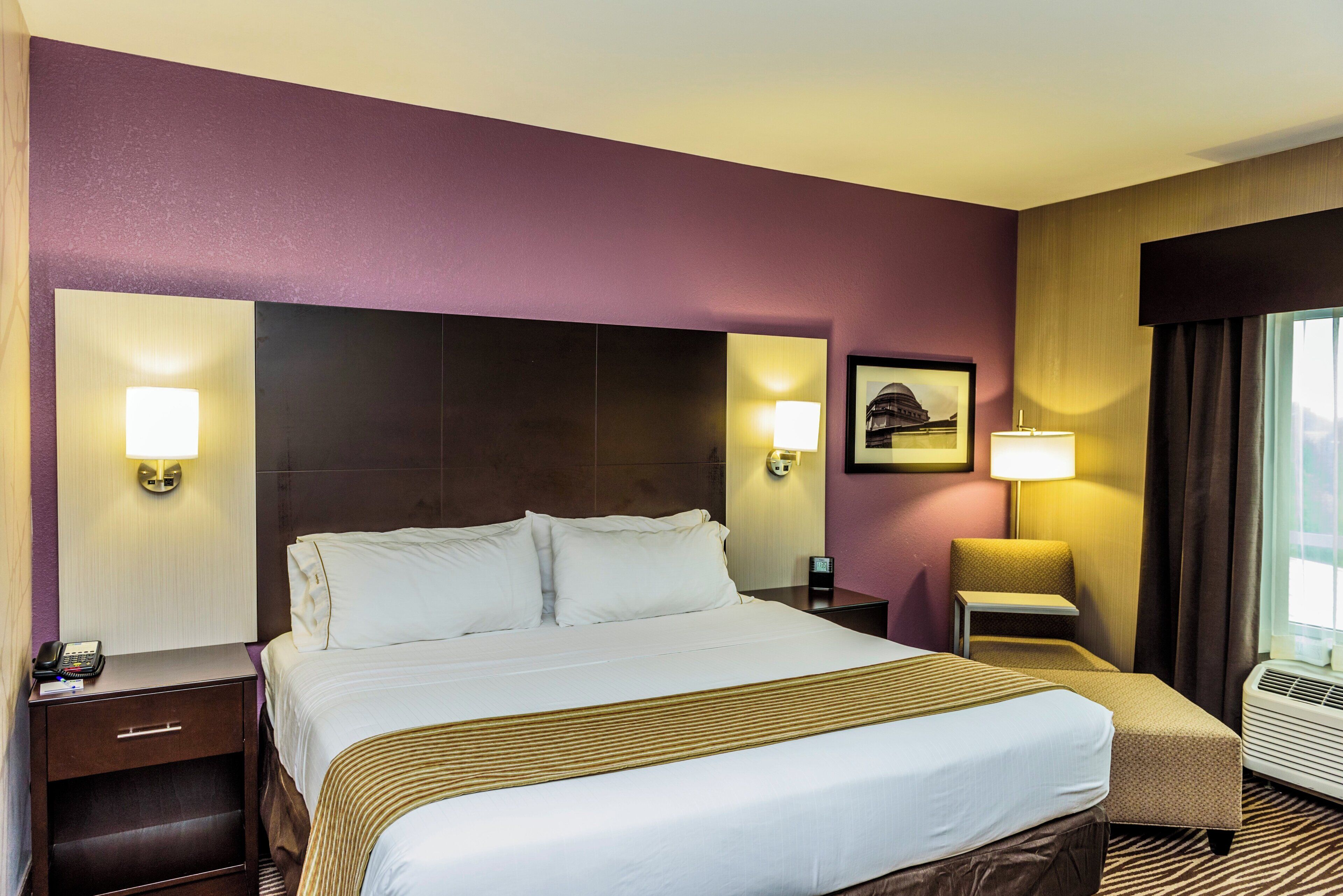 Una habitación de hotel con cama, mesitas de noche, una silla, una lámpara y un cuadro en la pared.