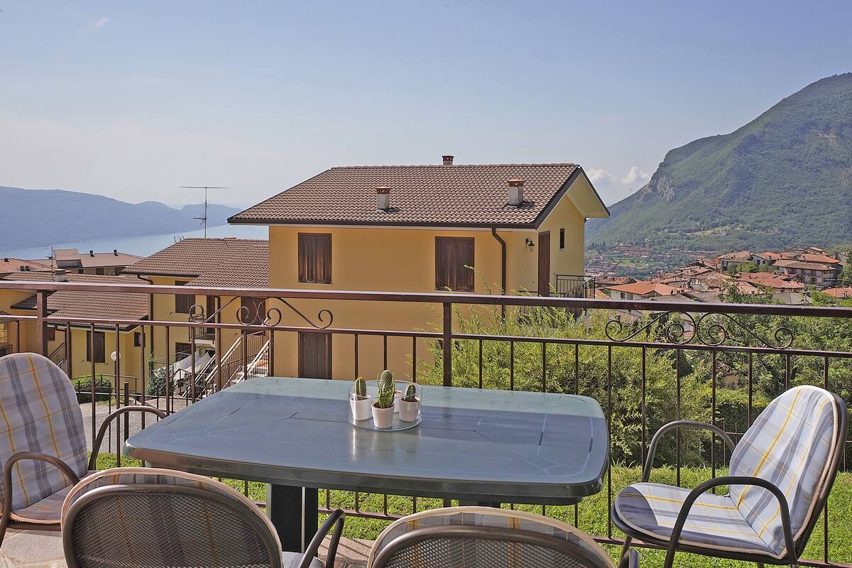 A balcony with a table and chairs, overlooking a mountainous landscape and a body of water.
