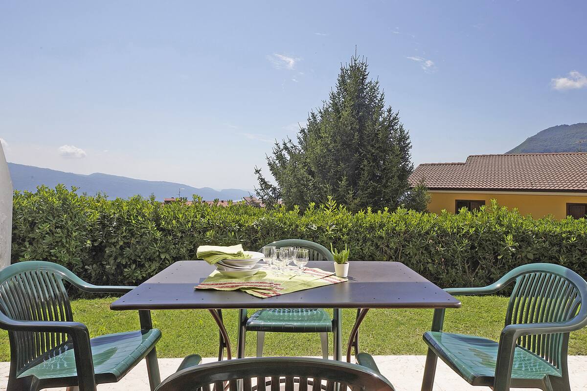 An outdoor dining area with a table set for two, surrounded by green chairs, against a backdrop of a hedge and a mountain.
