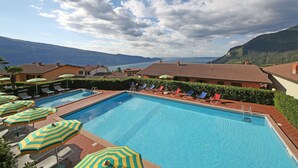 A swimming pool with multiple lounge chairs and umbrellas, surrounded by buildings with a view of a lake and mountains.