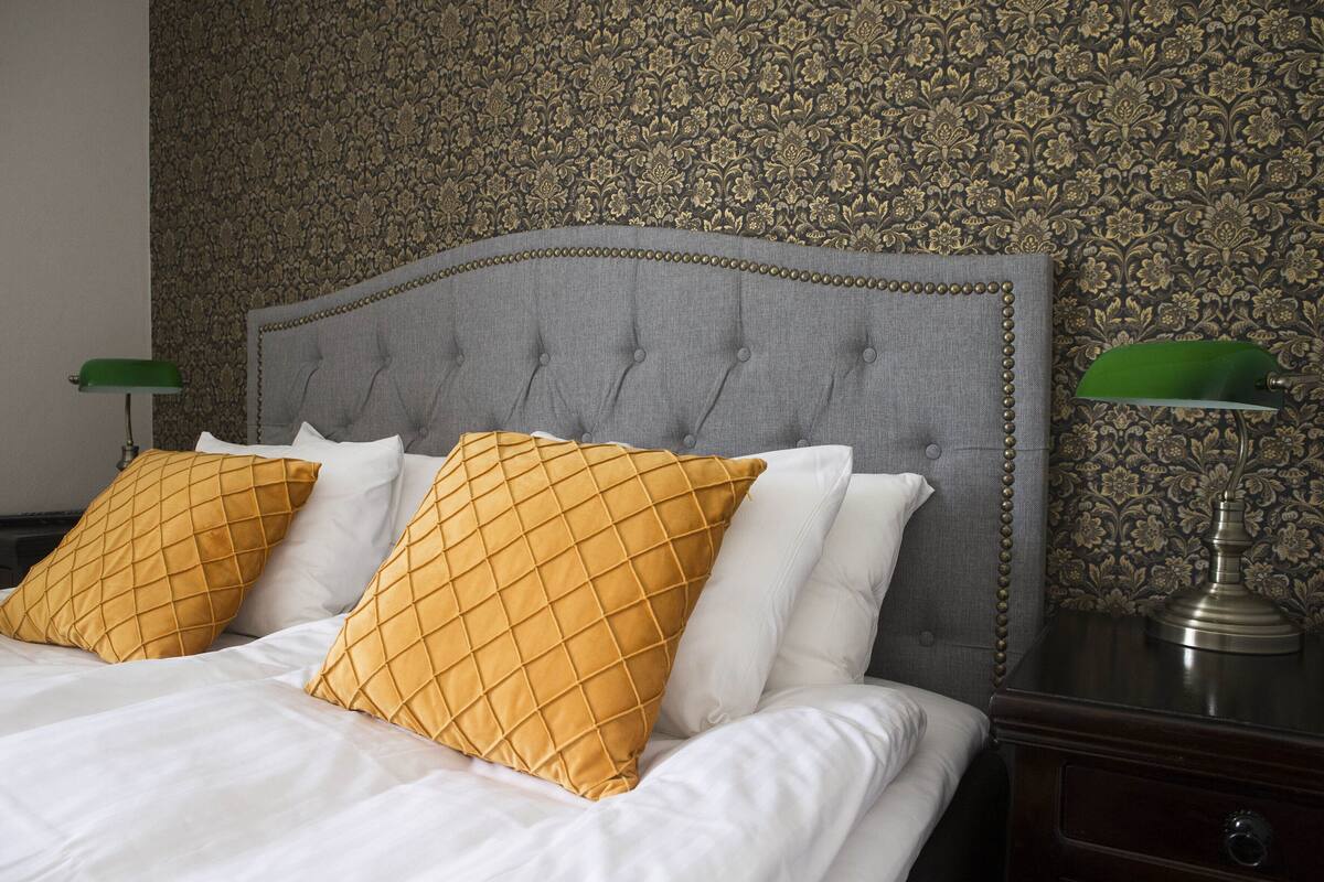 A bed with a grey upholstered headboard, two white pillows, and two mustard-colored pillows. There is a green desk lamp on a wooden nightstand beside the bed.