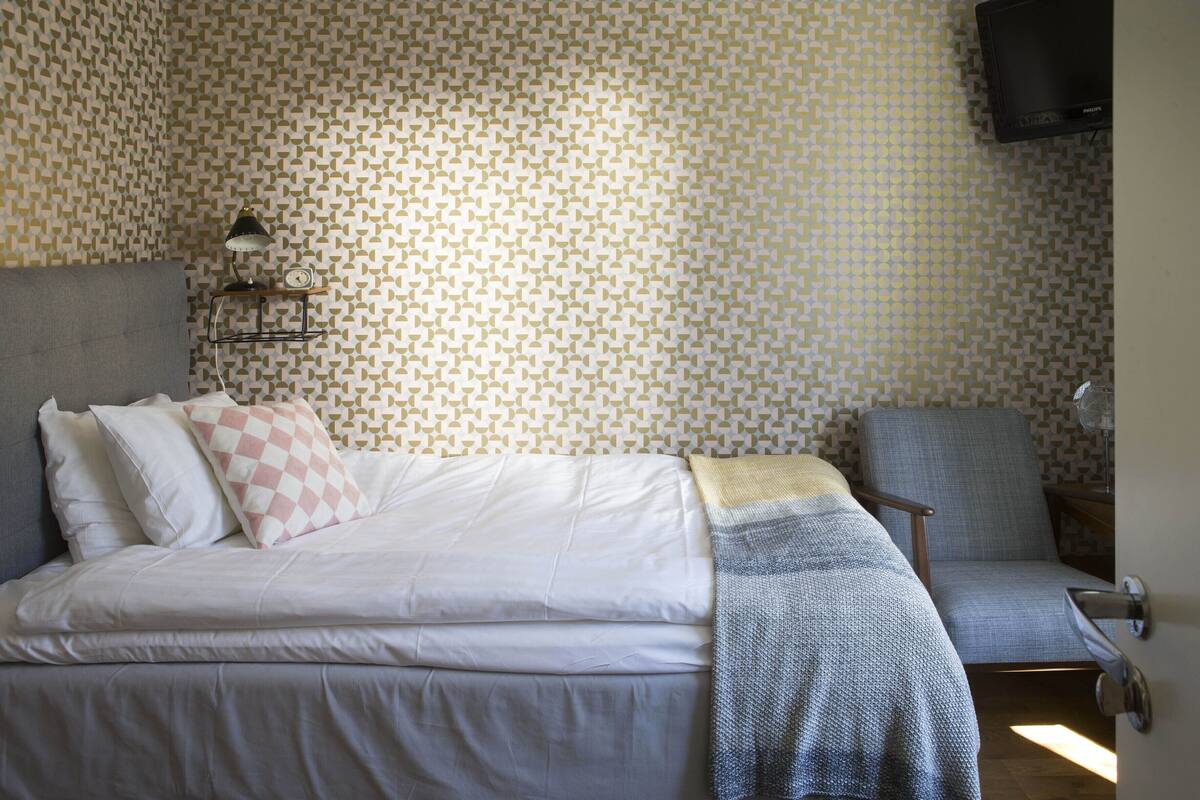A neatly made bed with pillows and a blanket, a chair, a shelf with a lamp and clock, and a patterned wall.