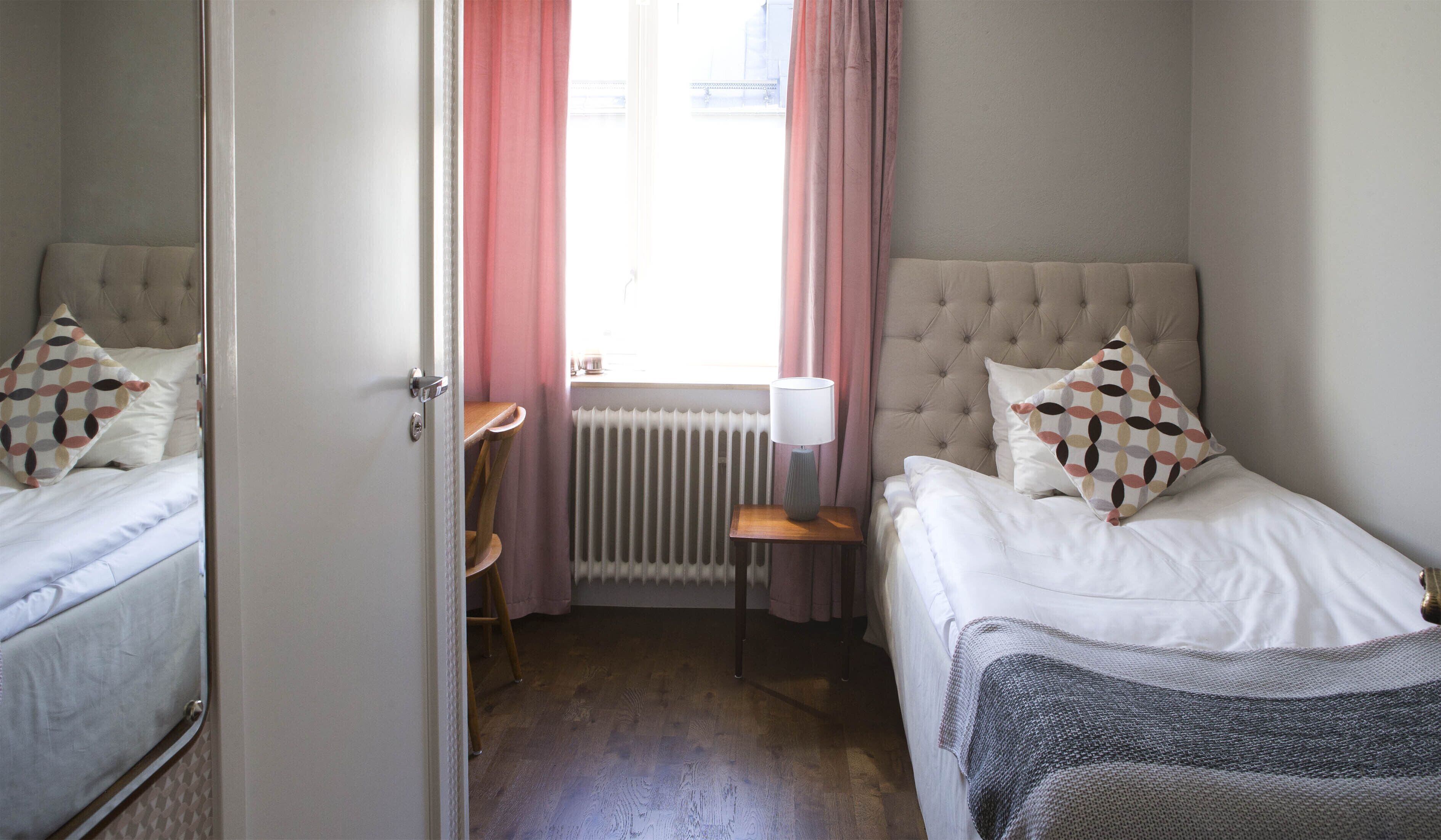 Un dormitorio con dos camas, una ventana con cortinas rosadas, un radiador y una mesita de noche con lámpara.