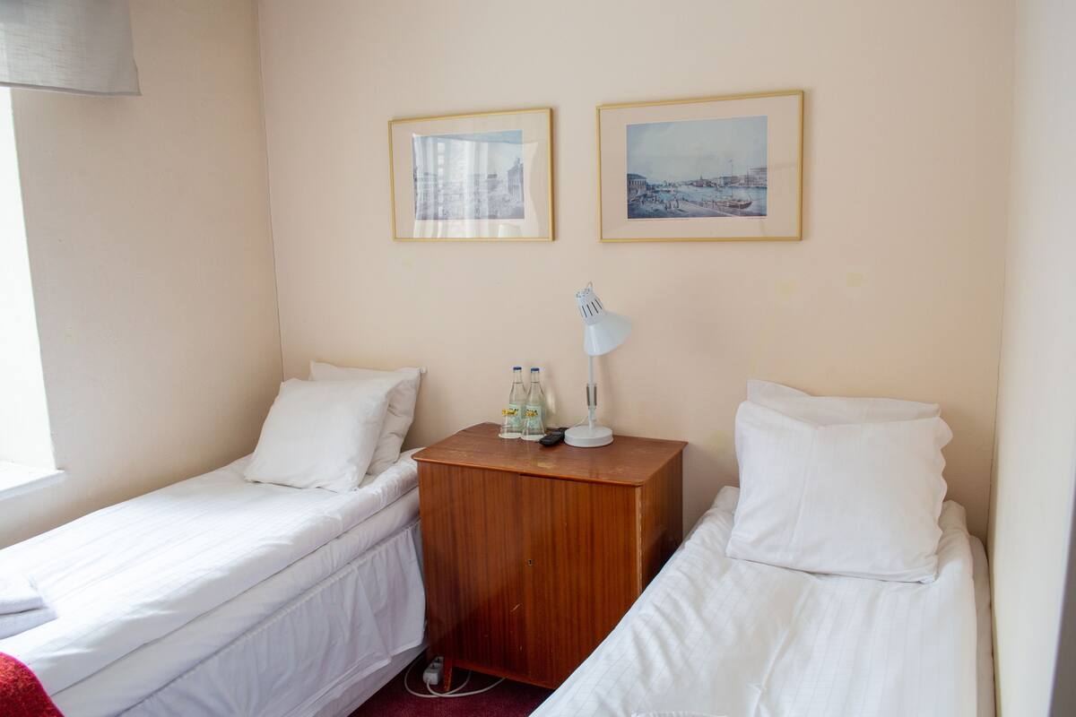 A small, clean room with two single beds, a wooden nightstand with a lamp, and two framed pictures on the wall.