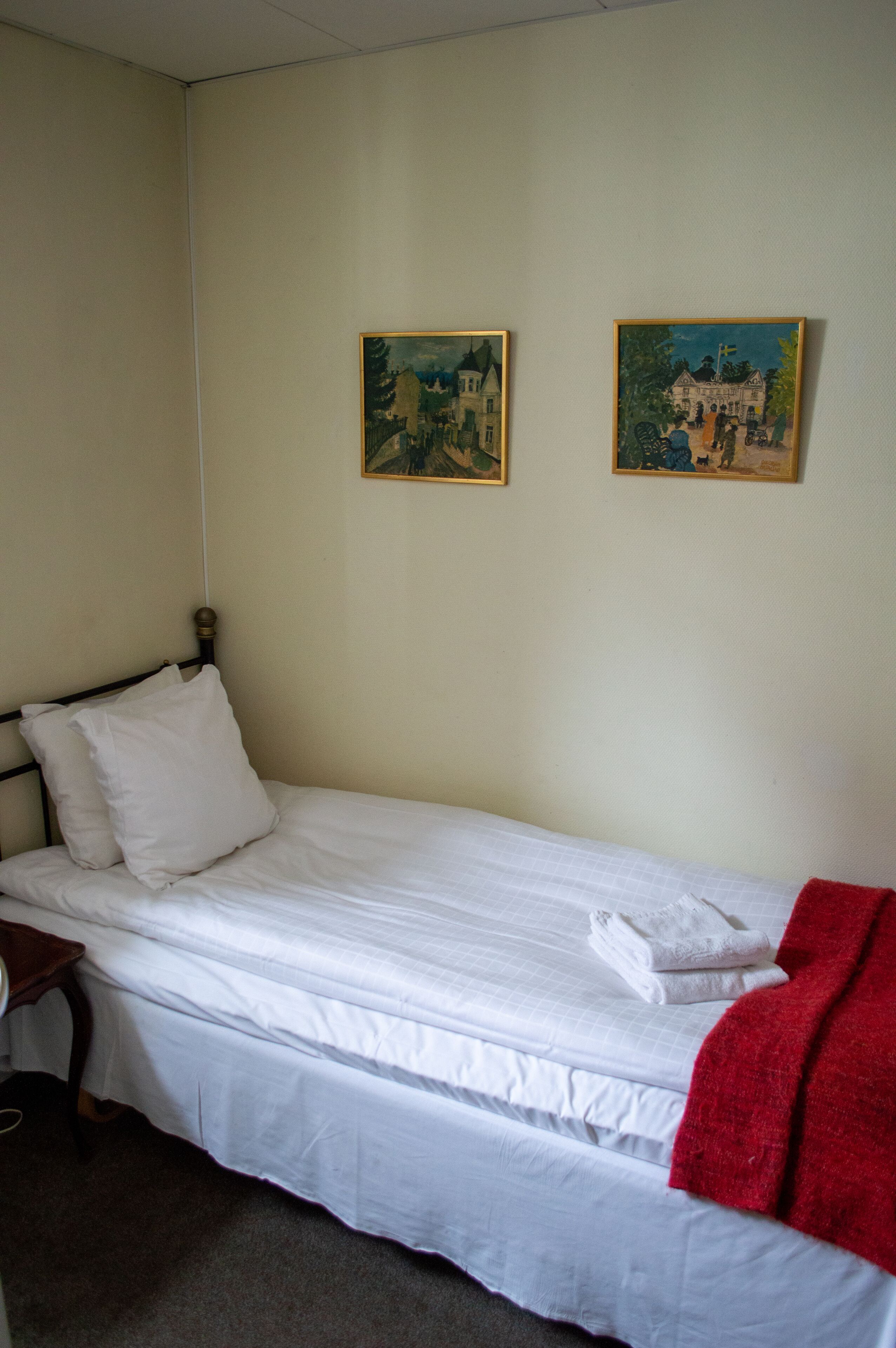 Un lit simple avec une literie blanche, une couverture rouge et deux tableaux accrochés au mur.