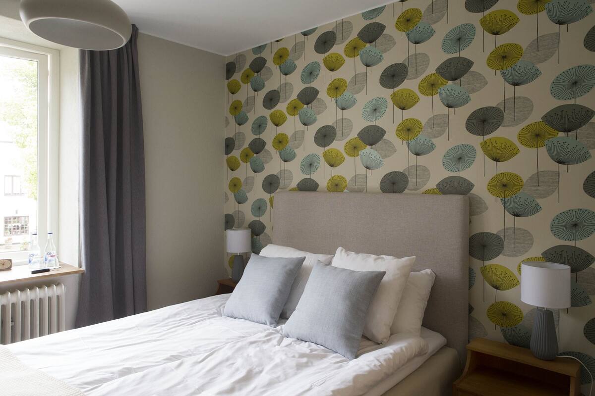 A neatly made bed with two pillows, a bedside table with a lamp, and a window with curtains.