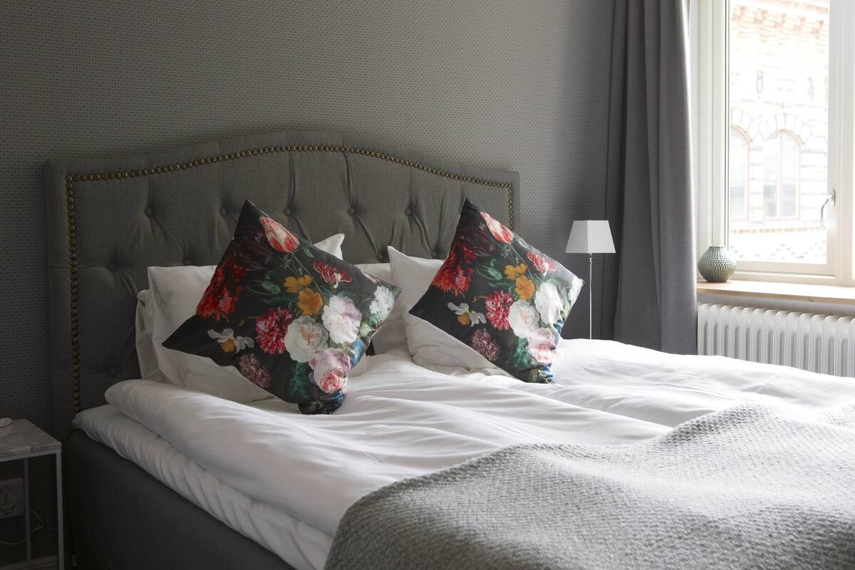 A neatly made bed with white linens and two floral-patterned pillows, a grey upholstered headboard, and a lamp on a bedside table.