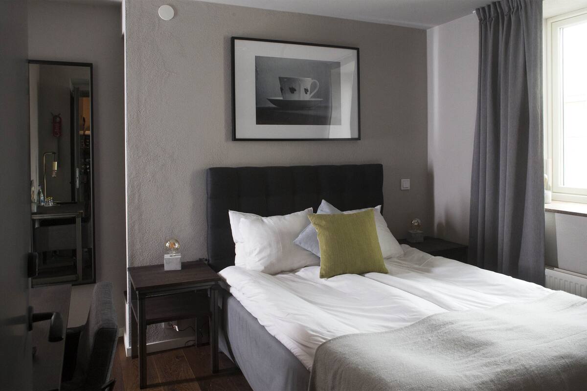 A hotel room with a neatly made bed, a bedside table with a lamp, a framed picture on the wall, and a window with curtains.