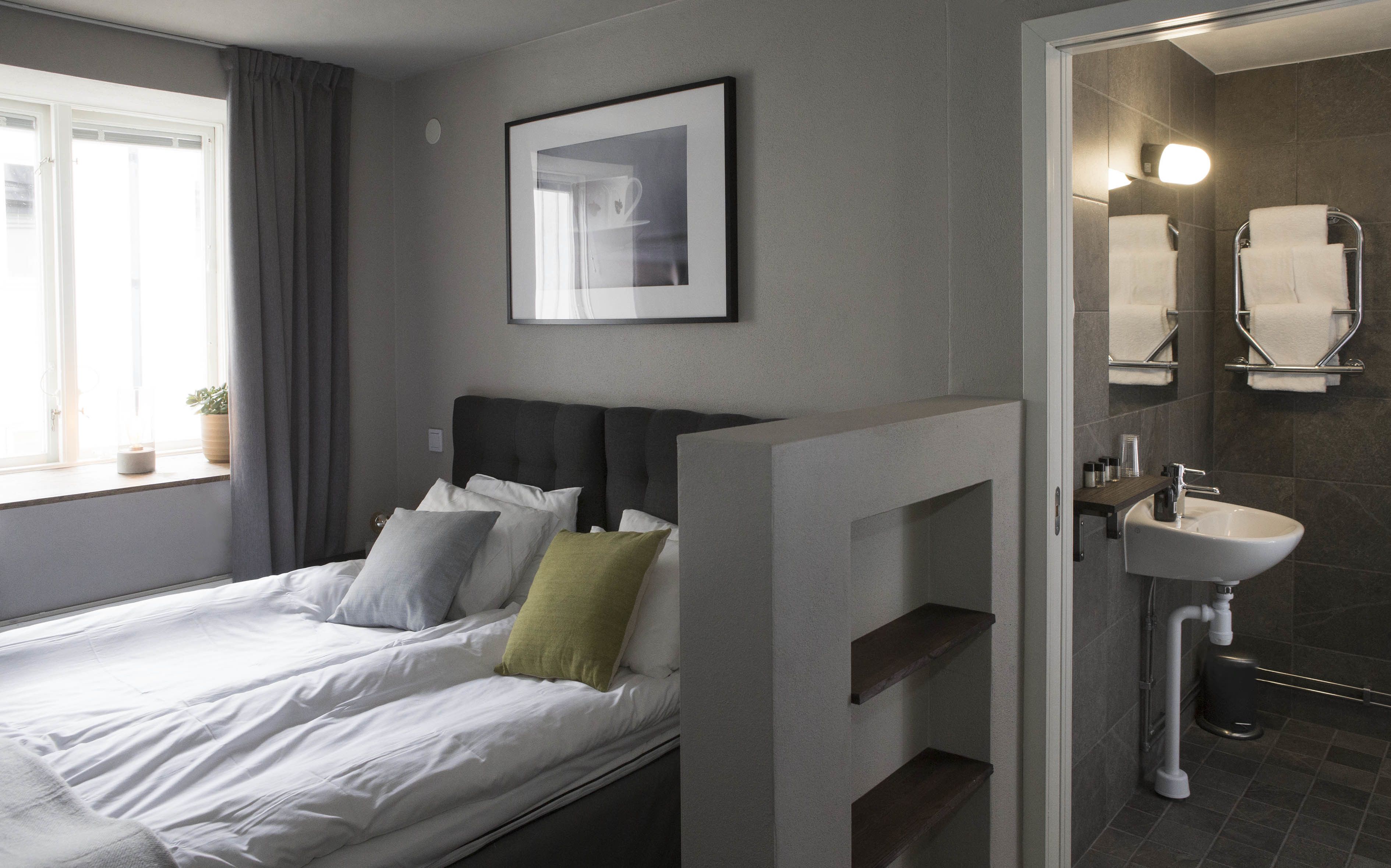 A modern hotel room with a bed, a bedside table, a wall-mounted picture, and a bathroom with a sink and mirror.
