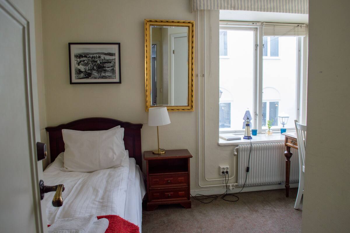 A hotel room with a bed, a nightstand, a lamp, a mirror, a window, and a radiator.