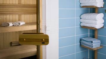 Un baño con paredes de azulejos azules, estantes de madera con toallas y artículos de aseo, y un banco de madera.