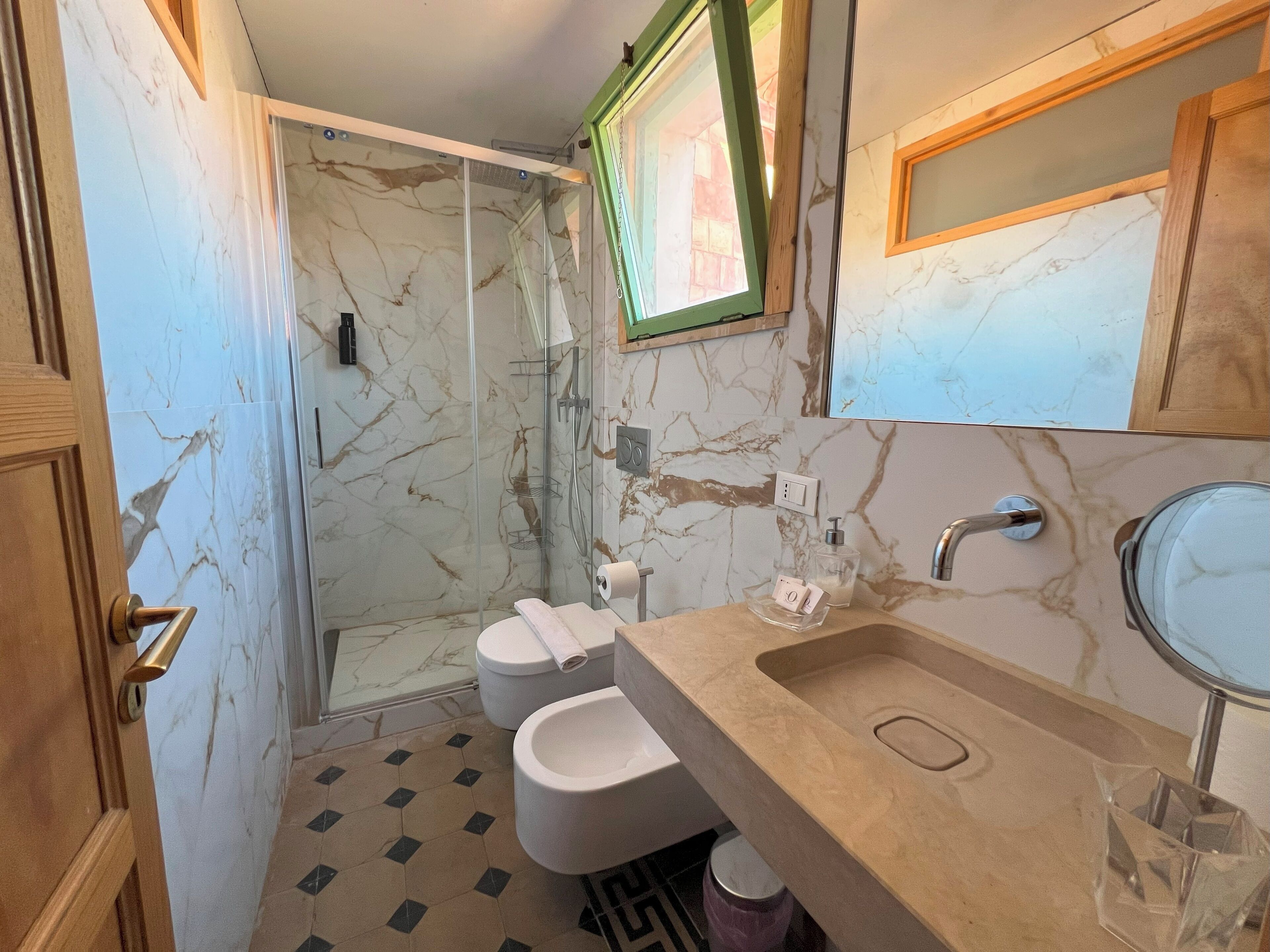 A bathroom with a marble wall, a shower, a toilet, and a sink with a mirror.