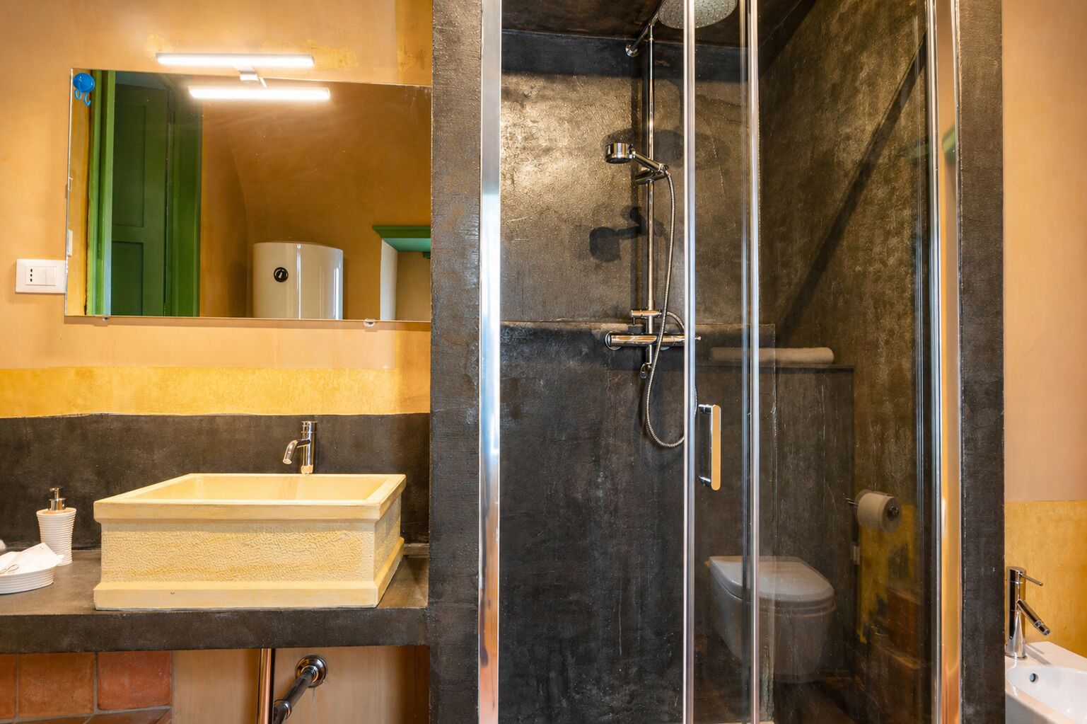 Ein Badezimmer mit einer gläsernen Duschkabine, einem Waschbecken mit Spiegel darüber und einer Toilette, die durch die Duschkabine sichtbar ist.