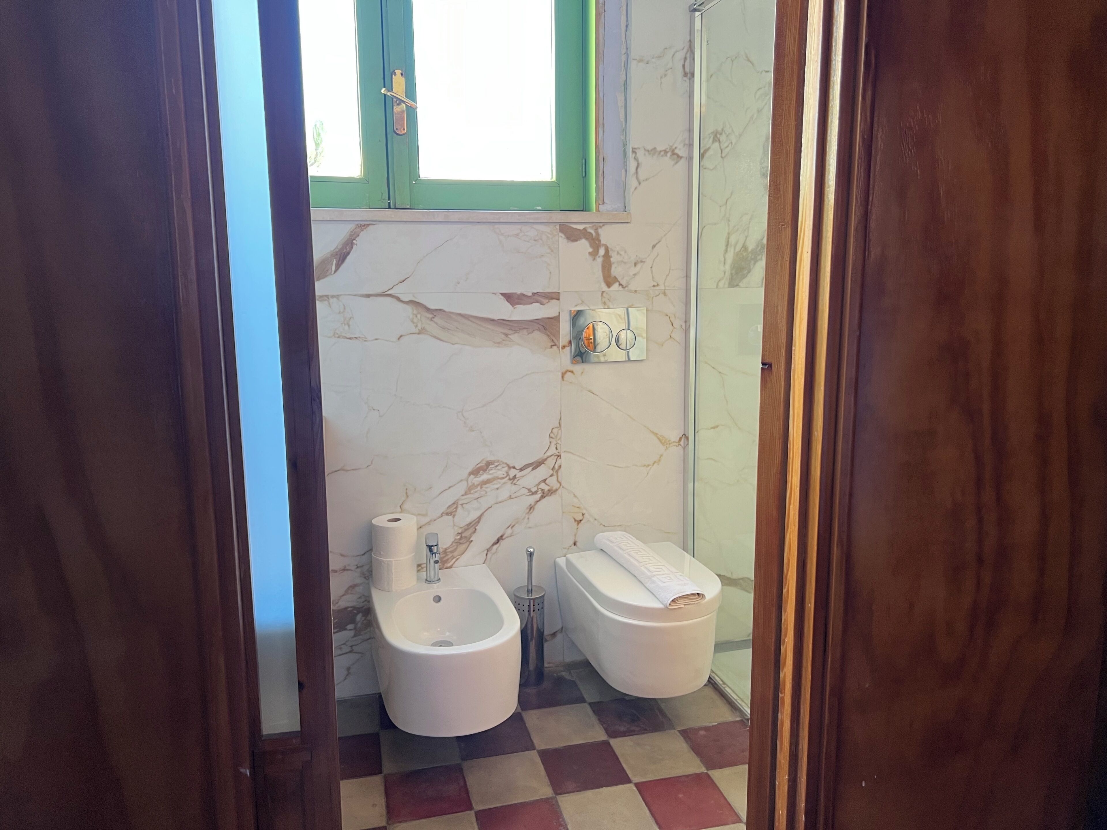 Un baño con bidé, inodoro, pared de mármol y una ventana con marco verde.