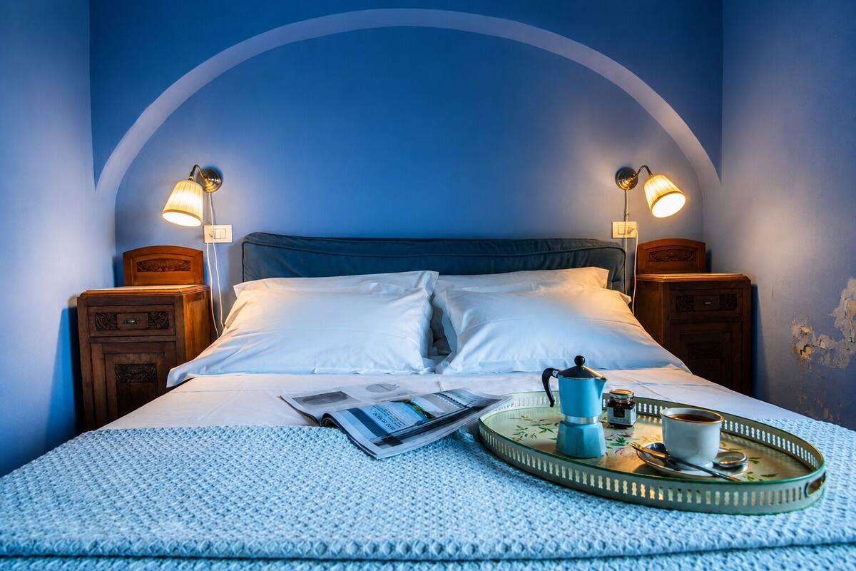 Un lit bien fait, avec du linge de lit blanc, deux lampes de chevet, un plateau avec une cafetière, une tasse et une cuillère, et un magazine posé sur le lit.