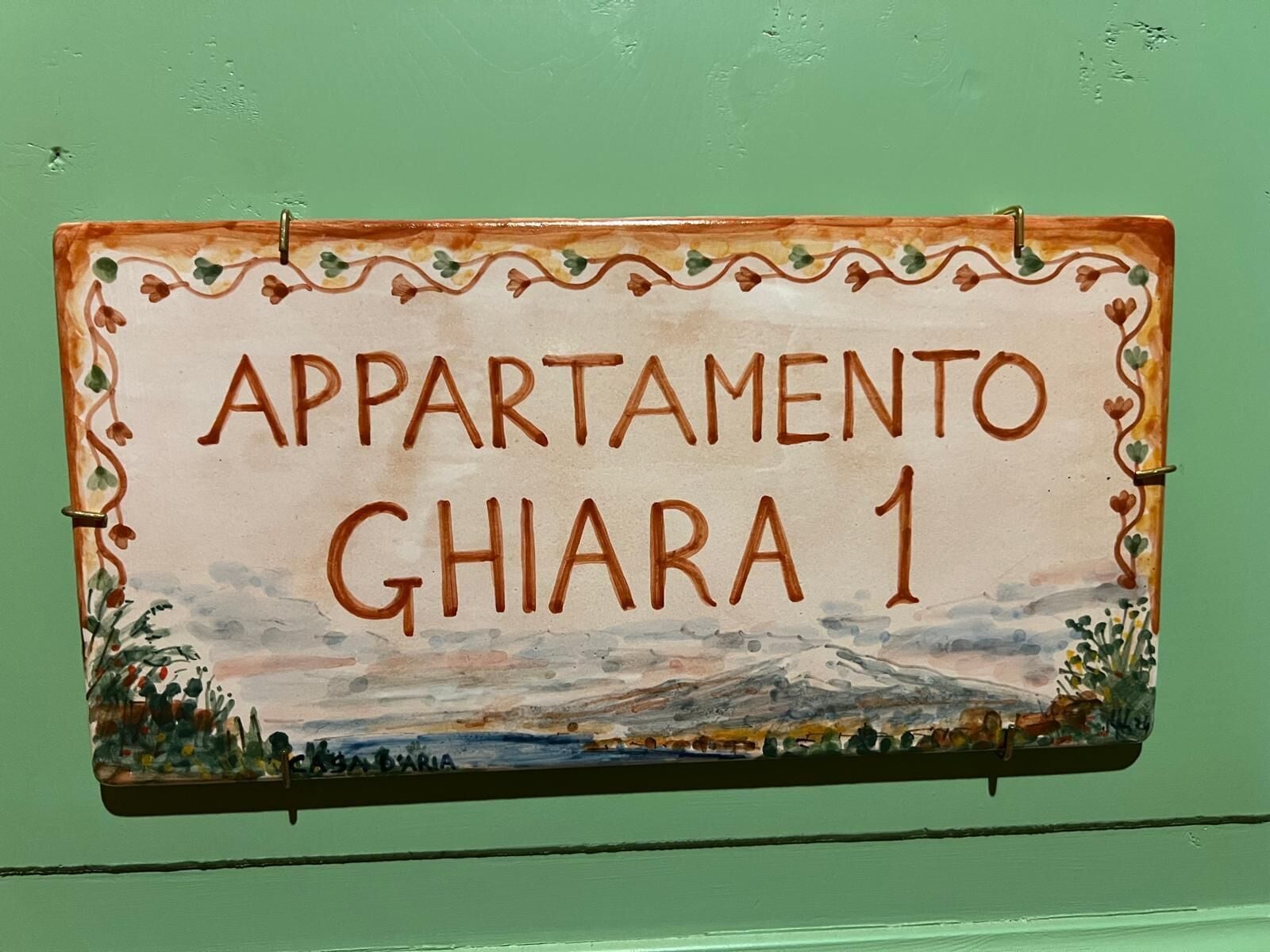 A sign with the text 'APARTAMENTO GHIARA 1' against a green wall, featuring a landscape painting.