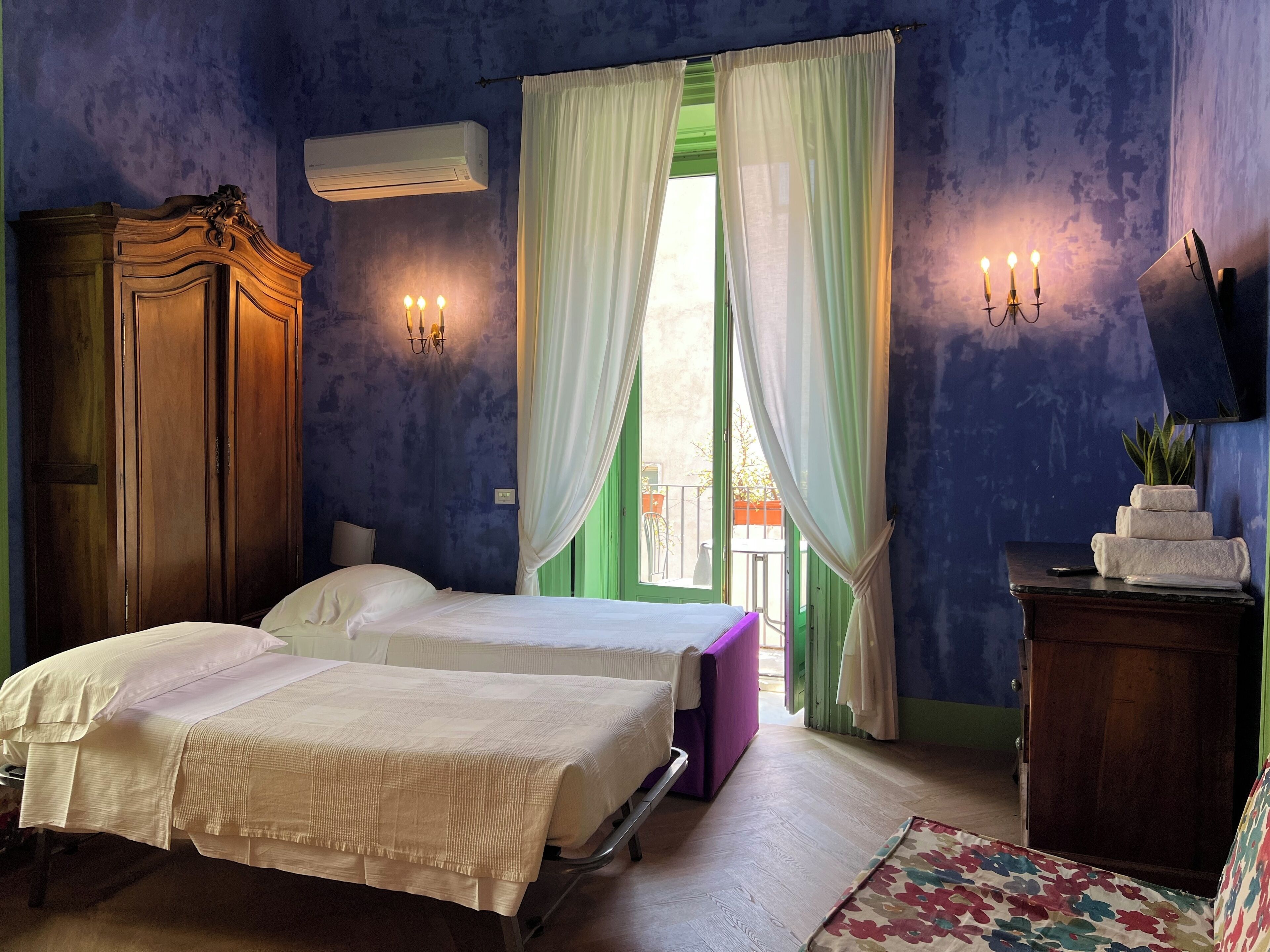 A room with two beds, a wooden wardrobe, a small table with a plant, and a window with curtains.