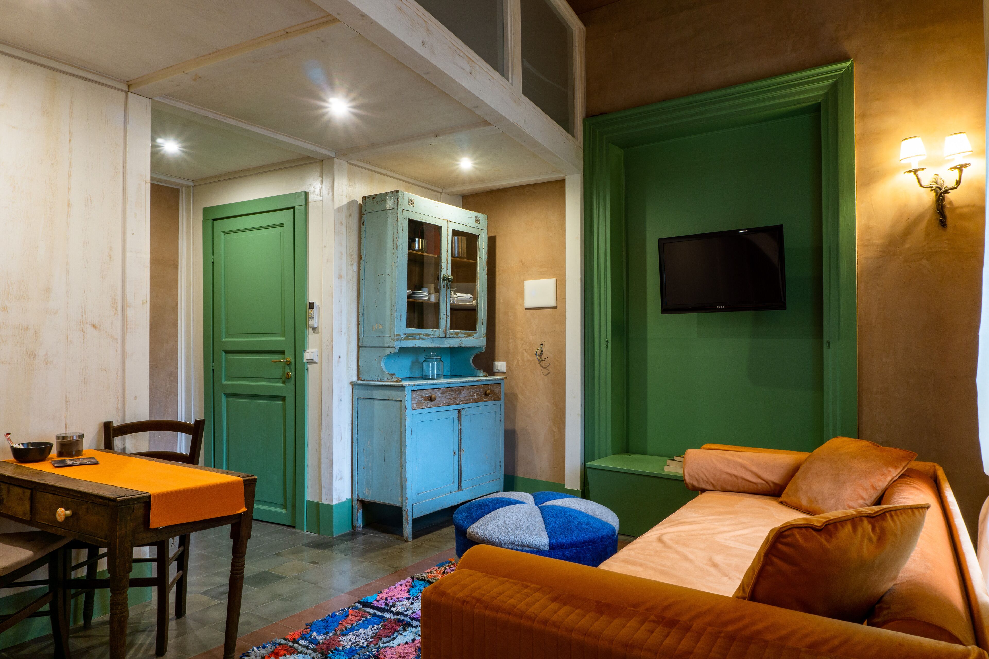 A room with a green door, a television mounted on the wall, a wooden table with an orange tablecloth, and a bed with orange bedding.