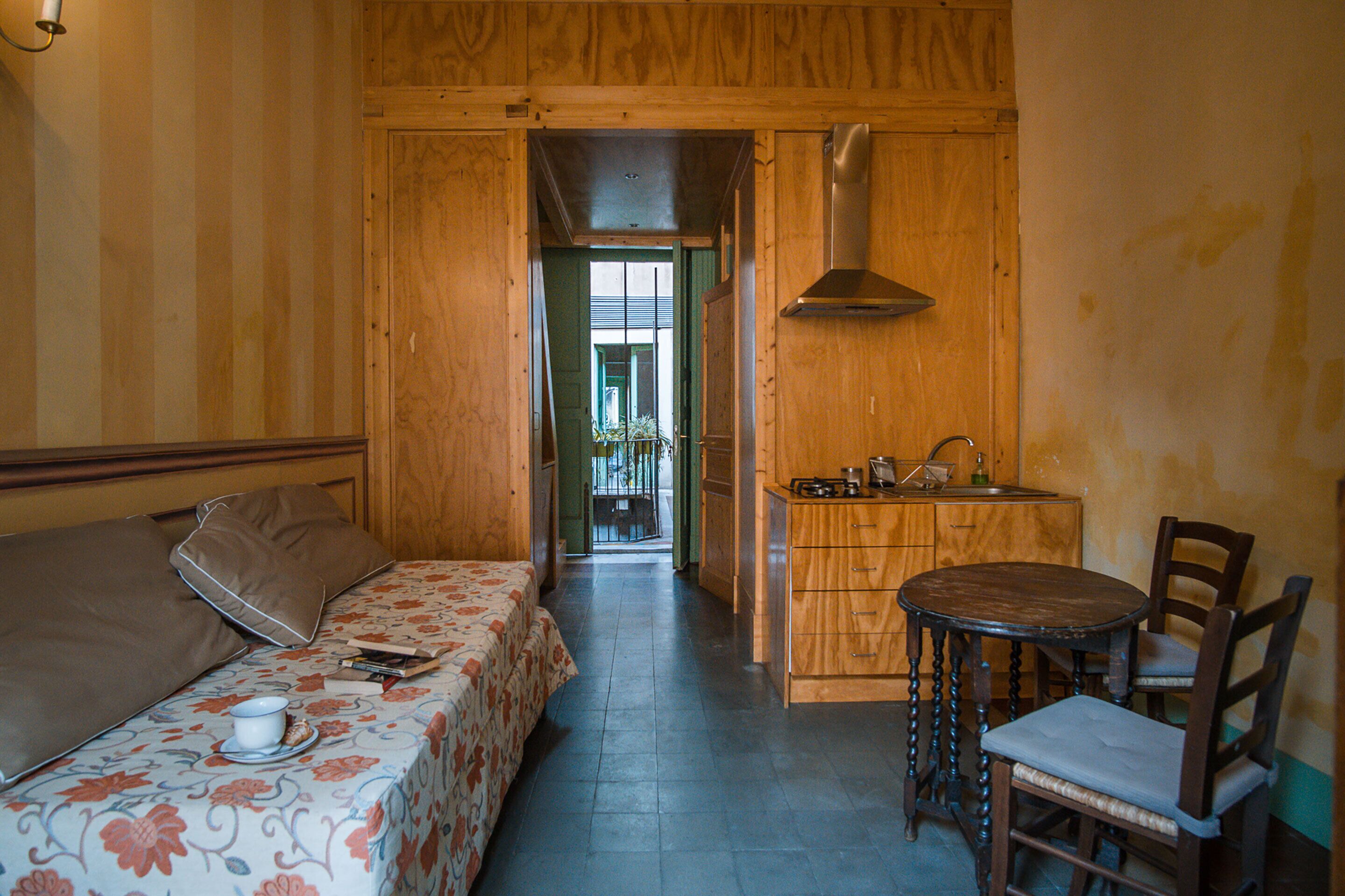 Una habitación con cama, una mesita con sillas, kitchenette y vistas a través de una puerta abierta.