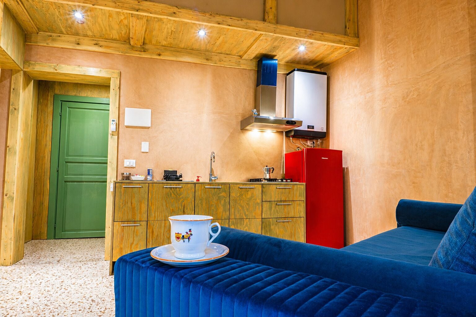 Una habitación compacta con kitchenette, una puerta verde, un sofá azul y un refrigerador rojo.