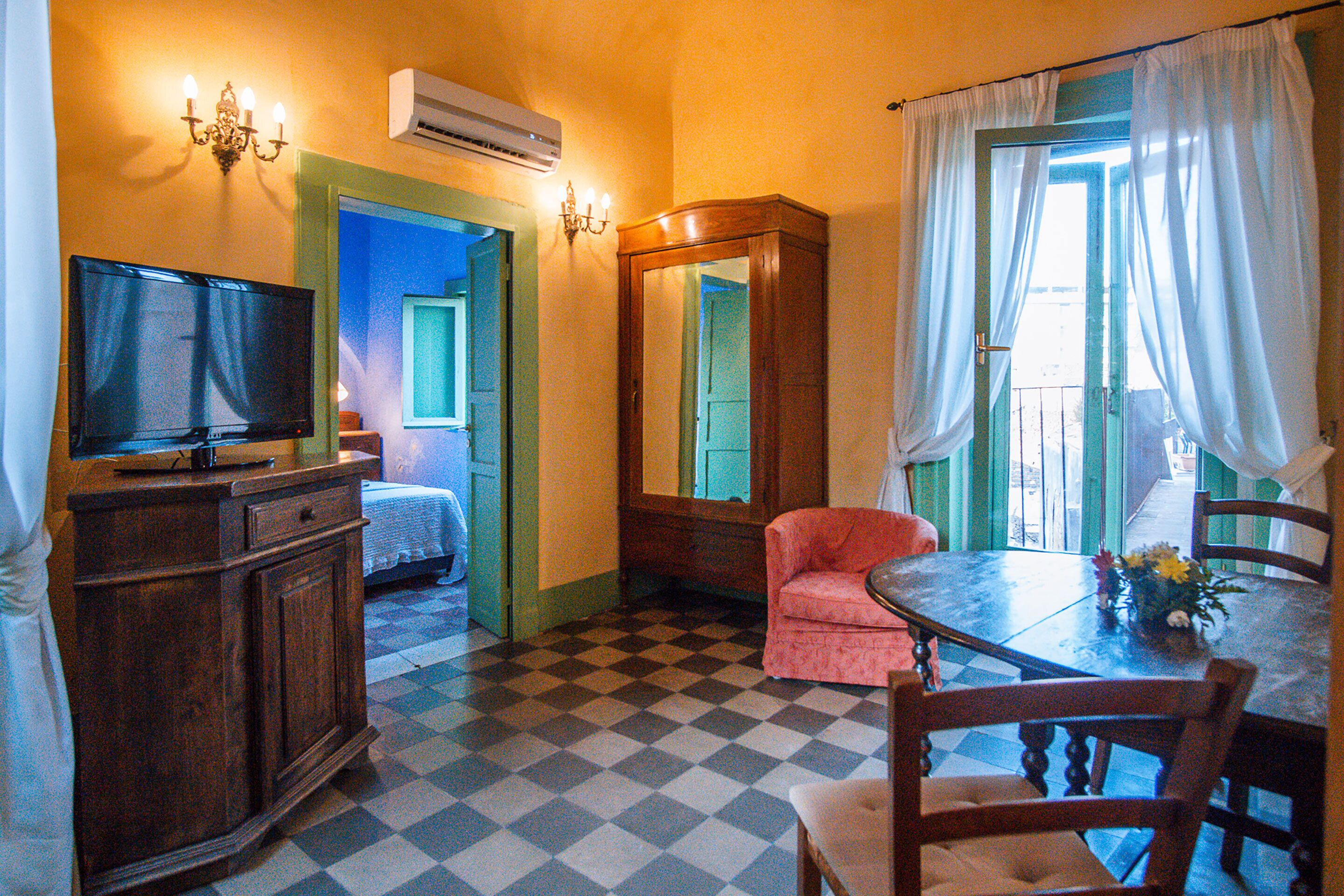 Una habitación de hotel con suelo a cuadros, un televisor, un aparador de madera, un espejo, una silla rosa, una mesa de comedor con sillas y una ventana con cortinas.