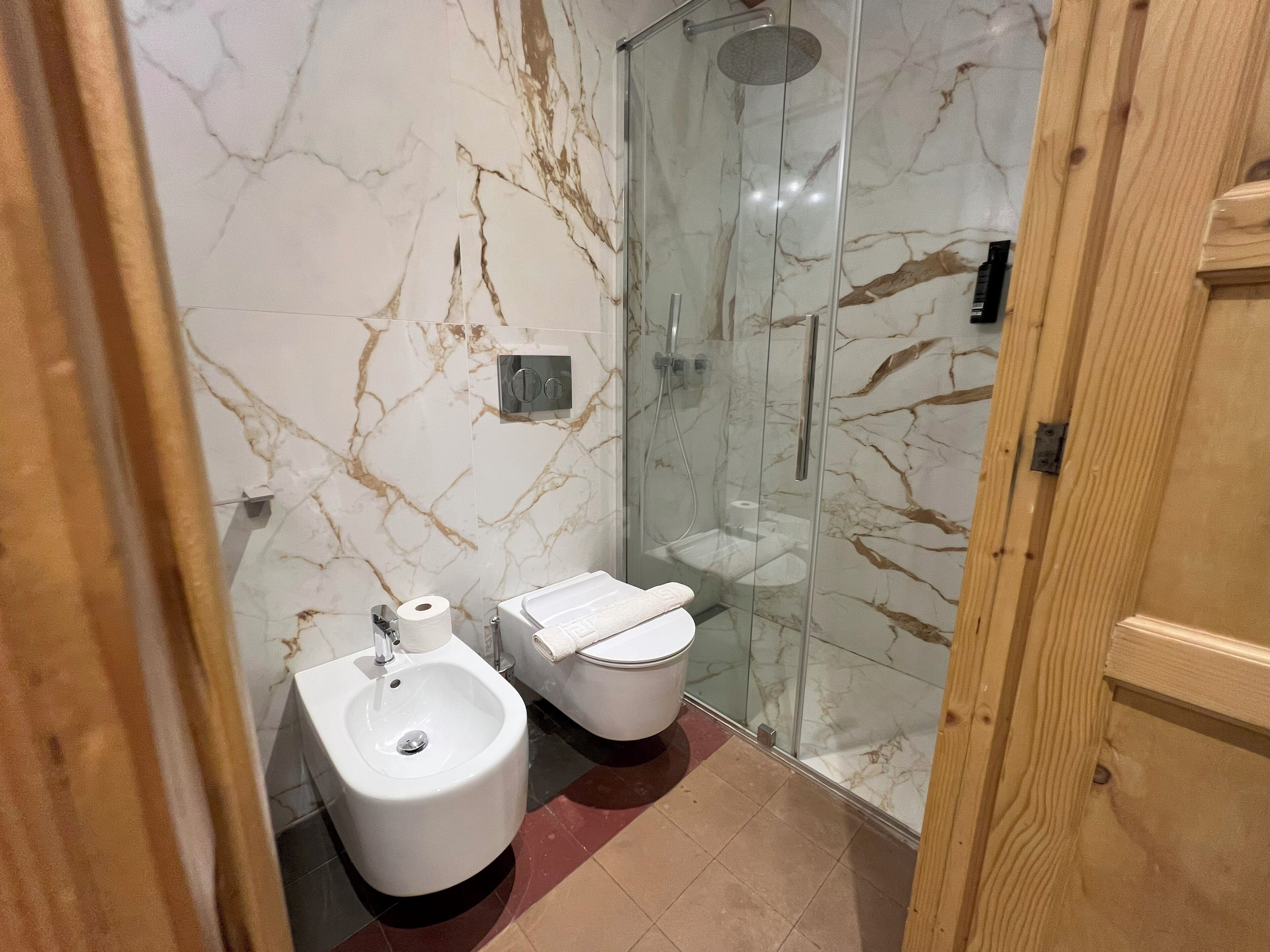 A bathroom with a marble wall, a bidet, a toilet, and a glass-enclosed shower.