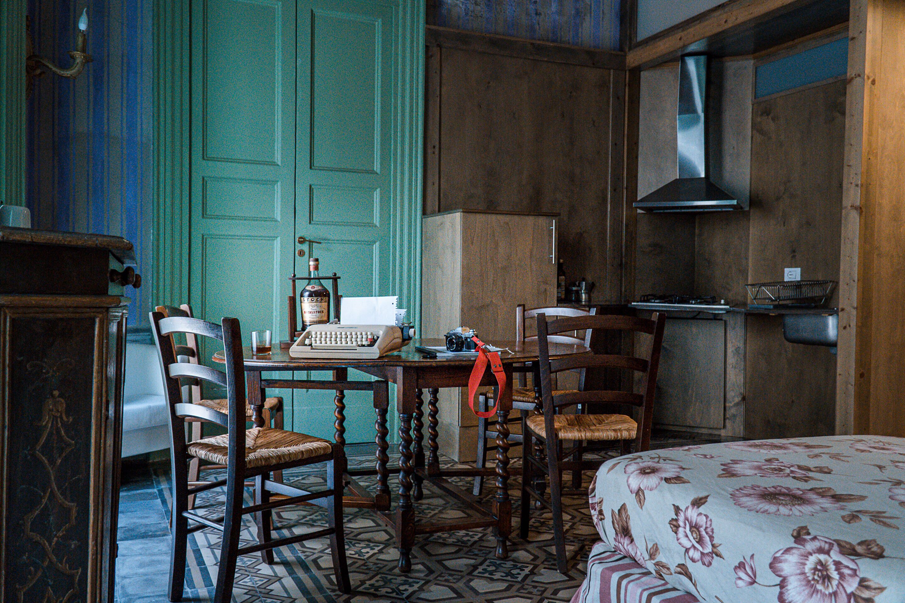A room with a wooden table, chairs, a bed with floral bedding, a vintage typewriter, a green door, and a kitchen area with a stove and sink.