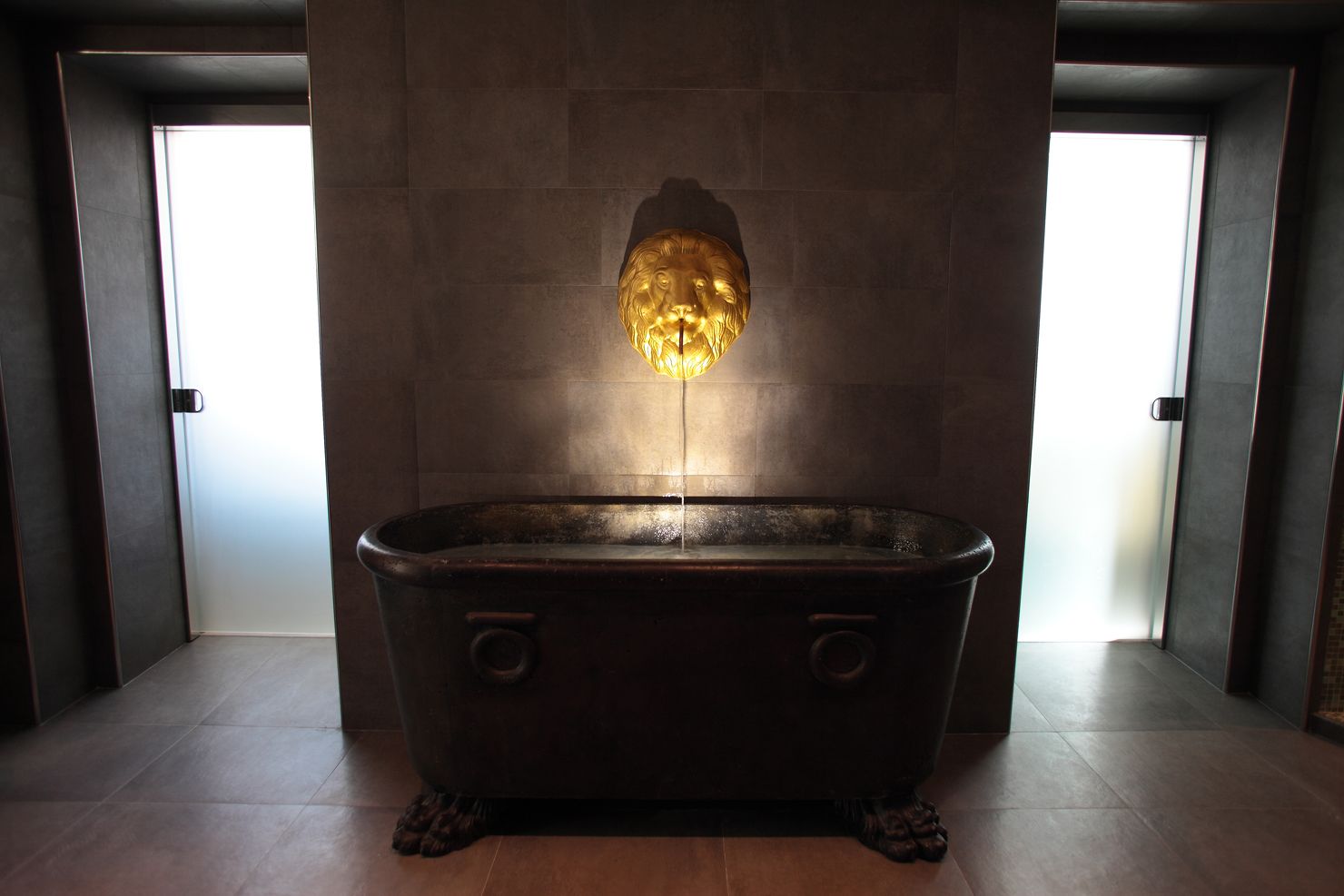 A freestanding bathtub in a modern bathroom with a dark floor and walls, and a large window on one side.